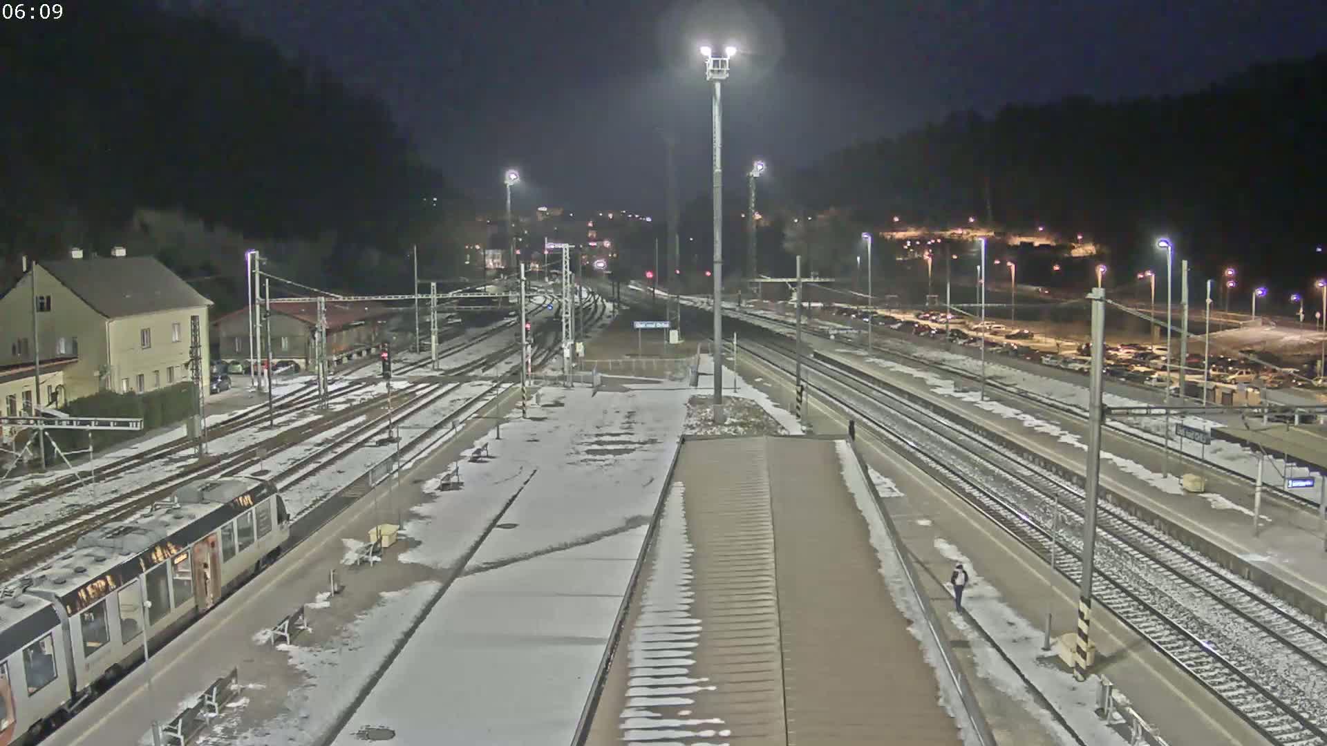 A train sits at a snow-dusted nighttime train station with multiple tracks and surrounding buildings.