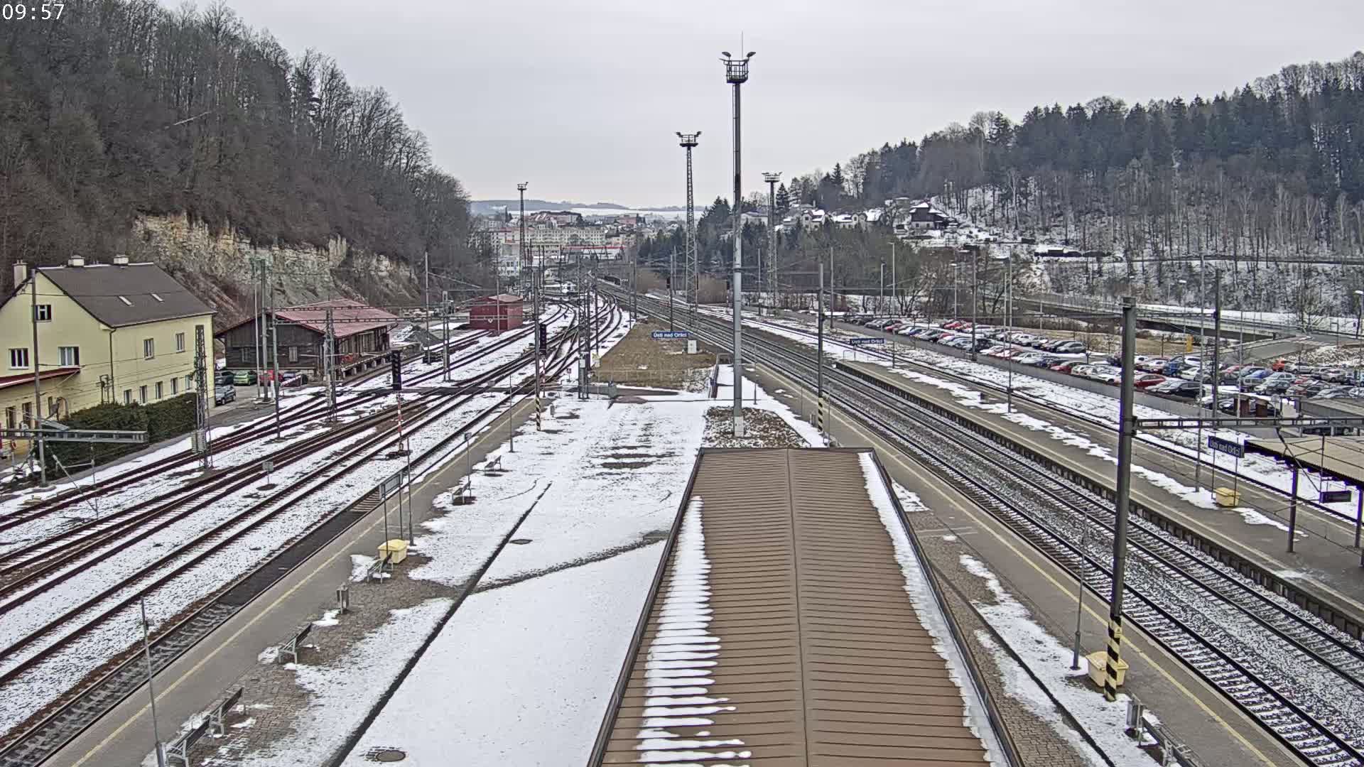 A snow-covered train station with multiple tracks and a large parking lot, surrounded by a wooded hillside under a cloudy sky.