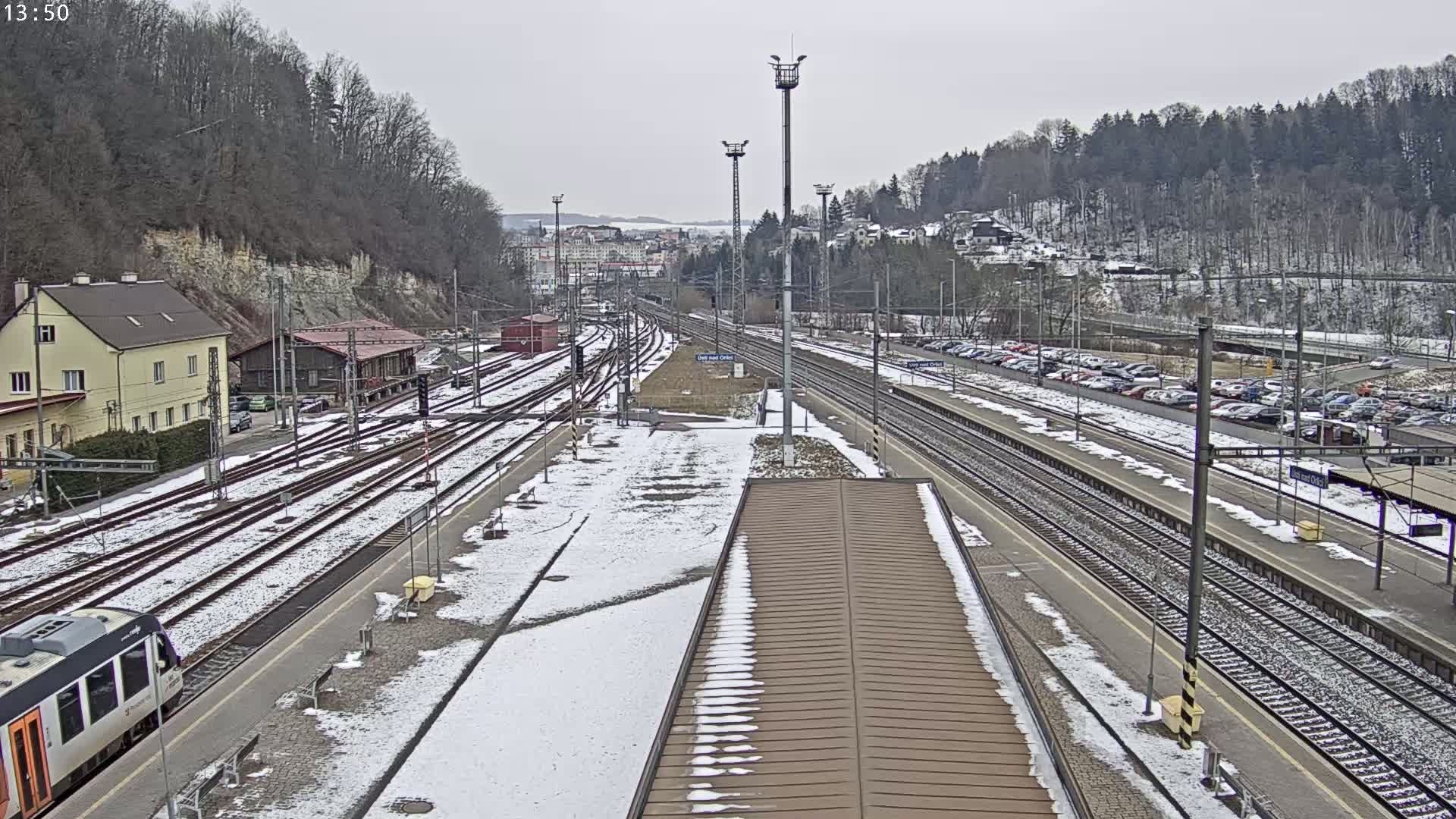 A snow-covered train station with multiple tracks, a train, and a parking lot is seen under an overcast sky.