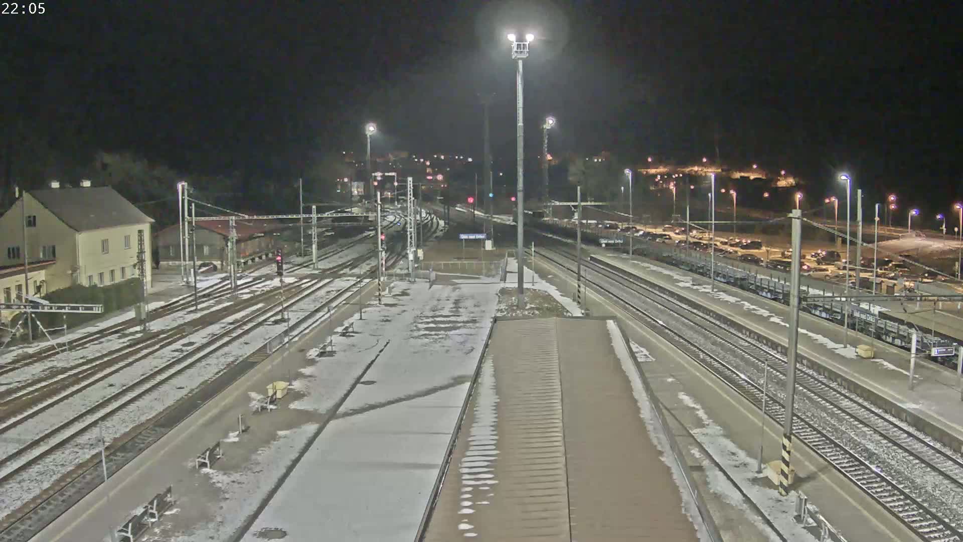 A nighttime view of a snow-dusted train station with multiple tracks and parked cars in the background under bright artificial lights.