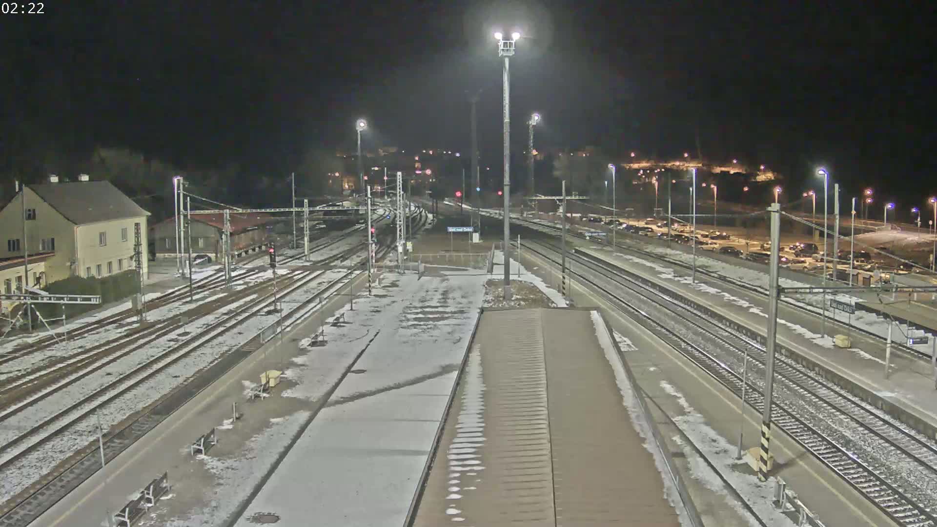 A snow-dusted train station at night, illuminated by bright lights, with multiple train tracks and a parking lot visible in the background.