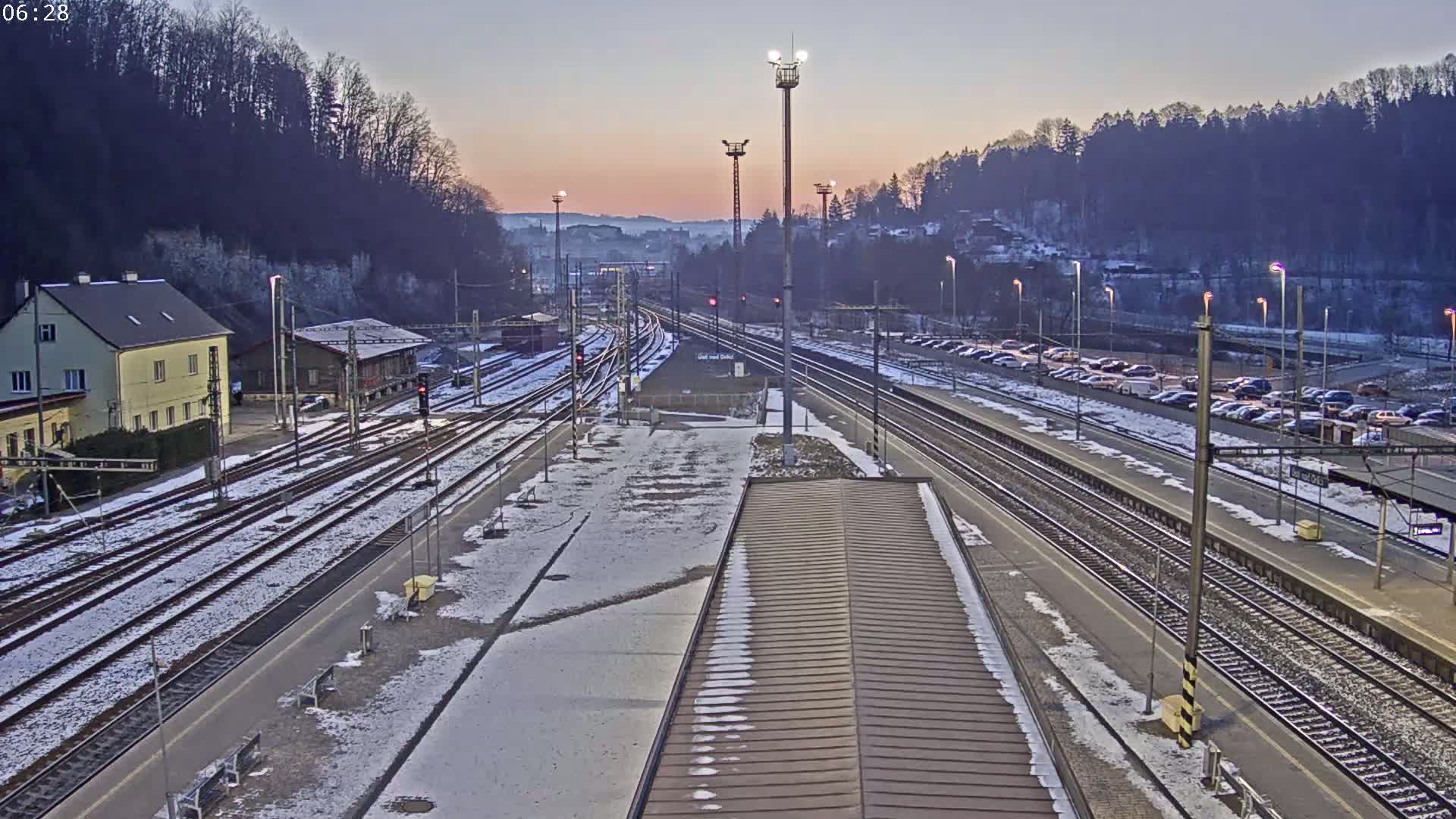 A snow-dusted train station with multiple tracks, a parking lot full of cars, and buildings under a twilight sky.