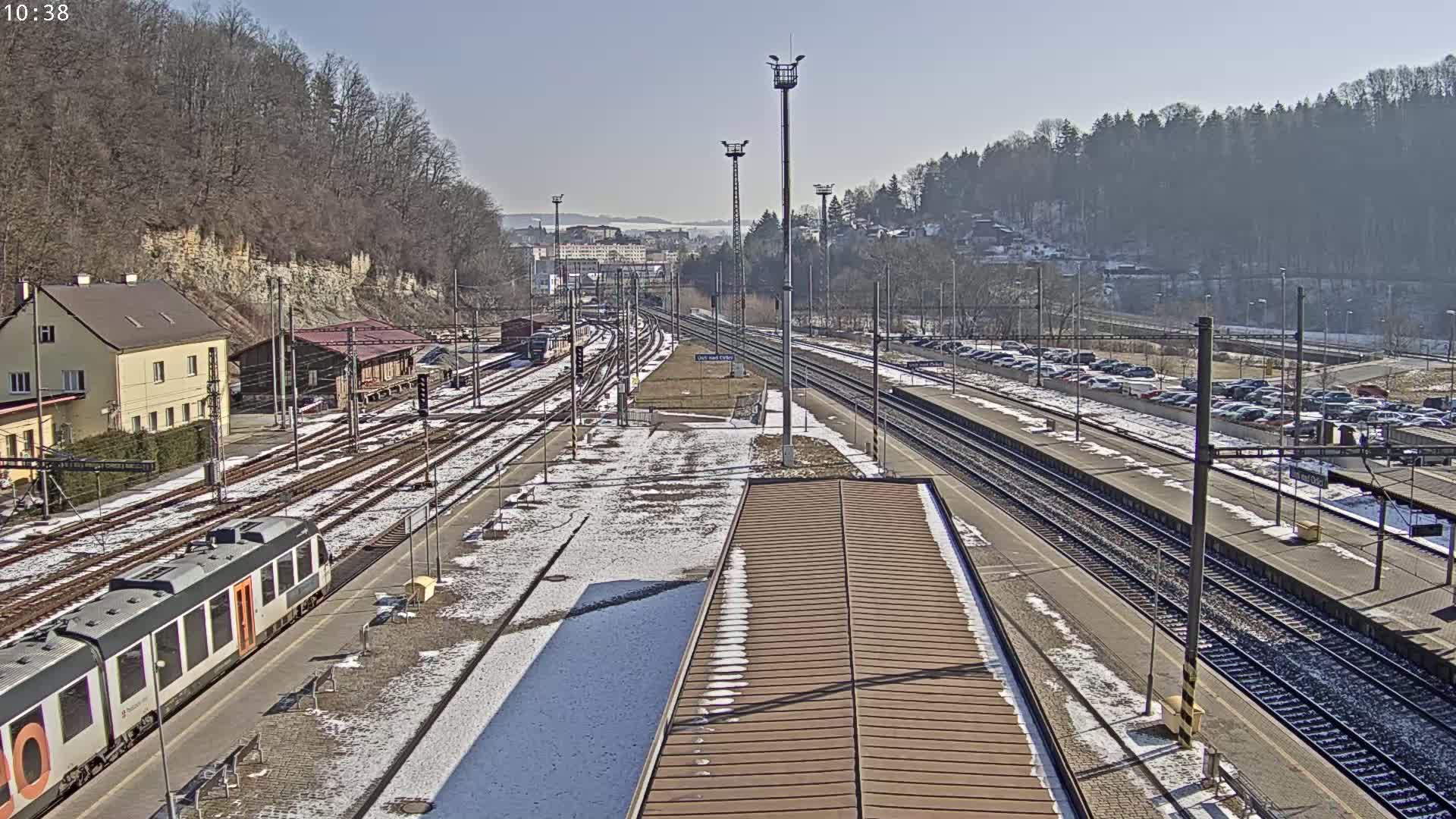 A train station with multiple train tracks, a partially snow-covered platform, and a large parking lot is visible on a sunny day.