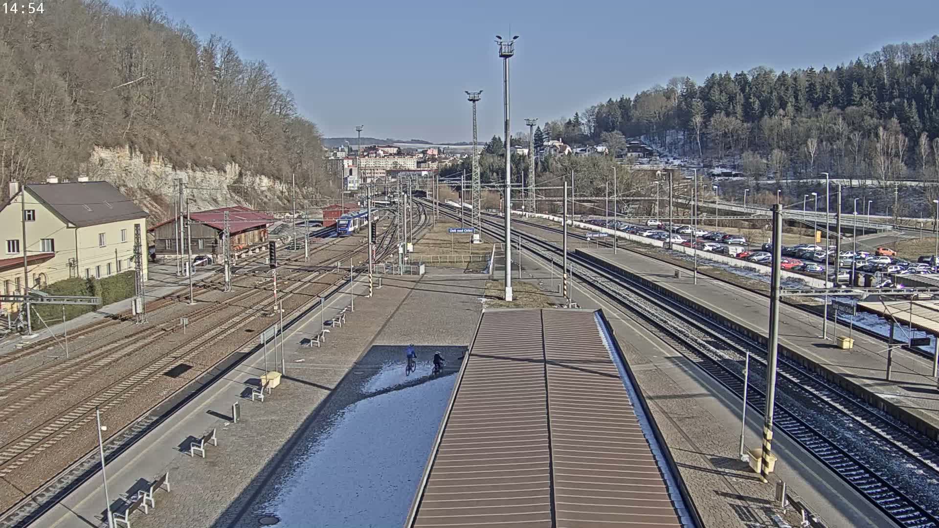 A sunny day at a train station with two cyclists riding near a partially snow-covered platform, multiple train tracks, and a large parking lot visible beyond the station.