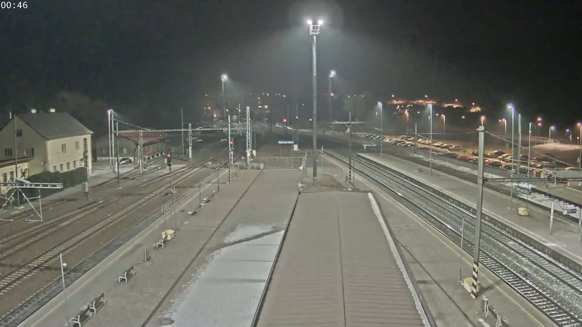 A nighttime view of a train station with multiple tracks, platforms, and a parking lot, under slightly hazy conditions.