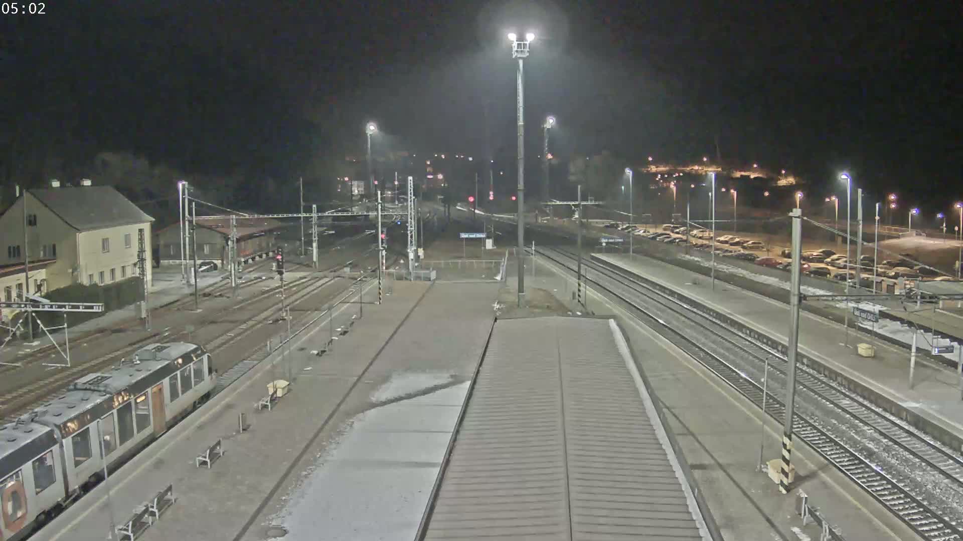 A nighttime view of a train station with a train on a platform, multiple tracks, and a parking lot with cars, some snow or ice is on the ground.