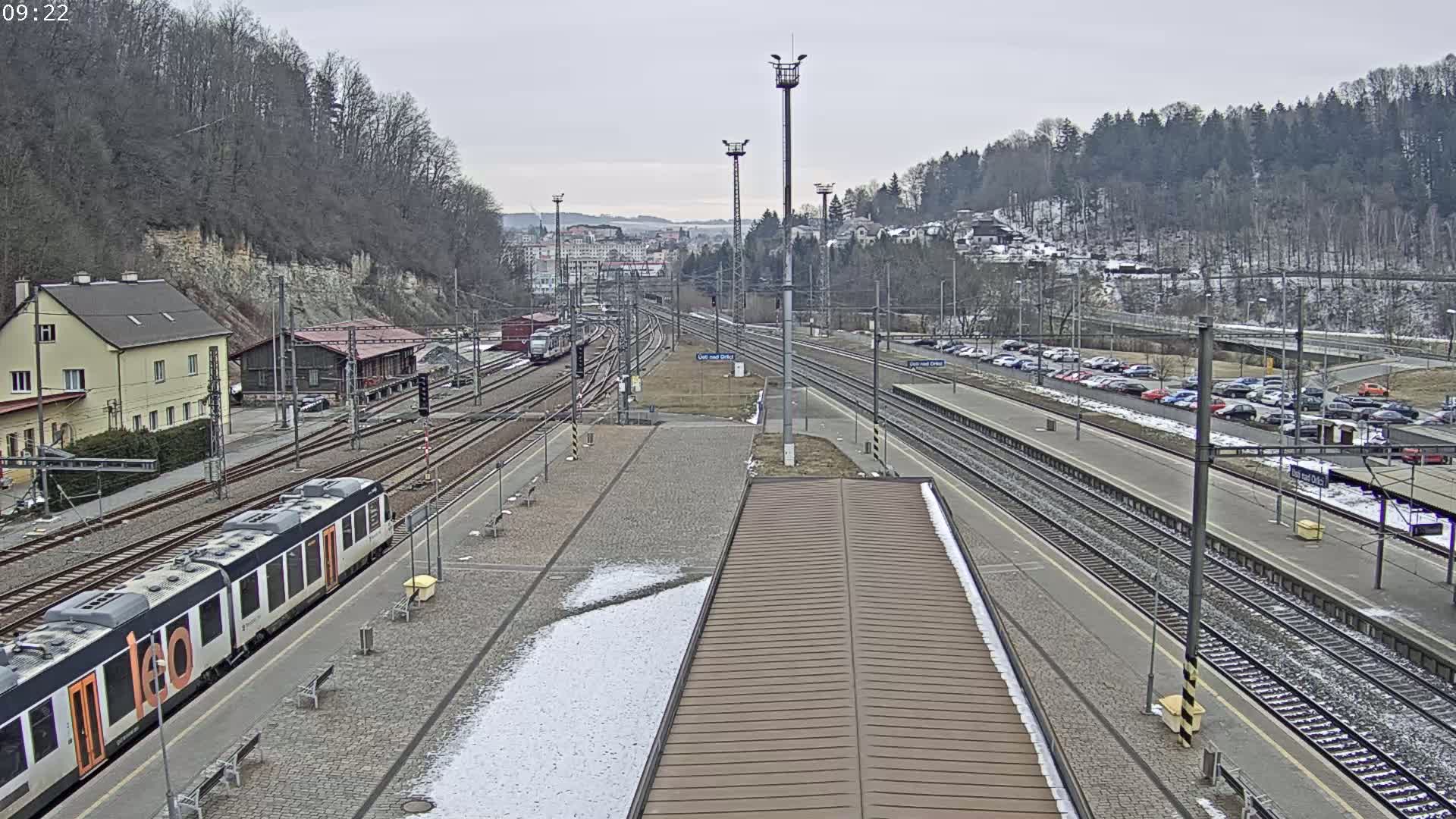 A train station with multiple tracks, a train on one of the tracks, and cars parked in a nearby lot on an overcast day with some snow on the ground.