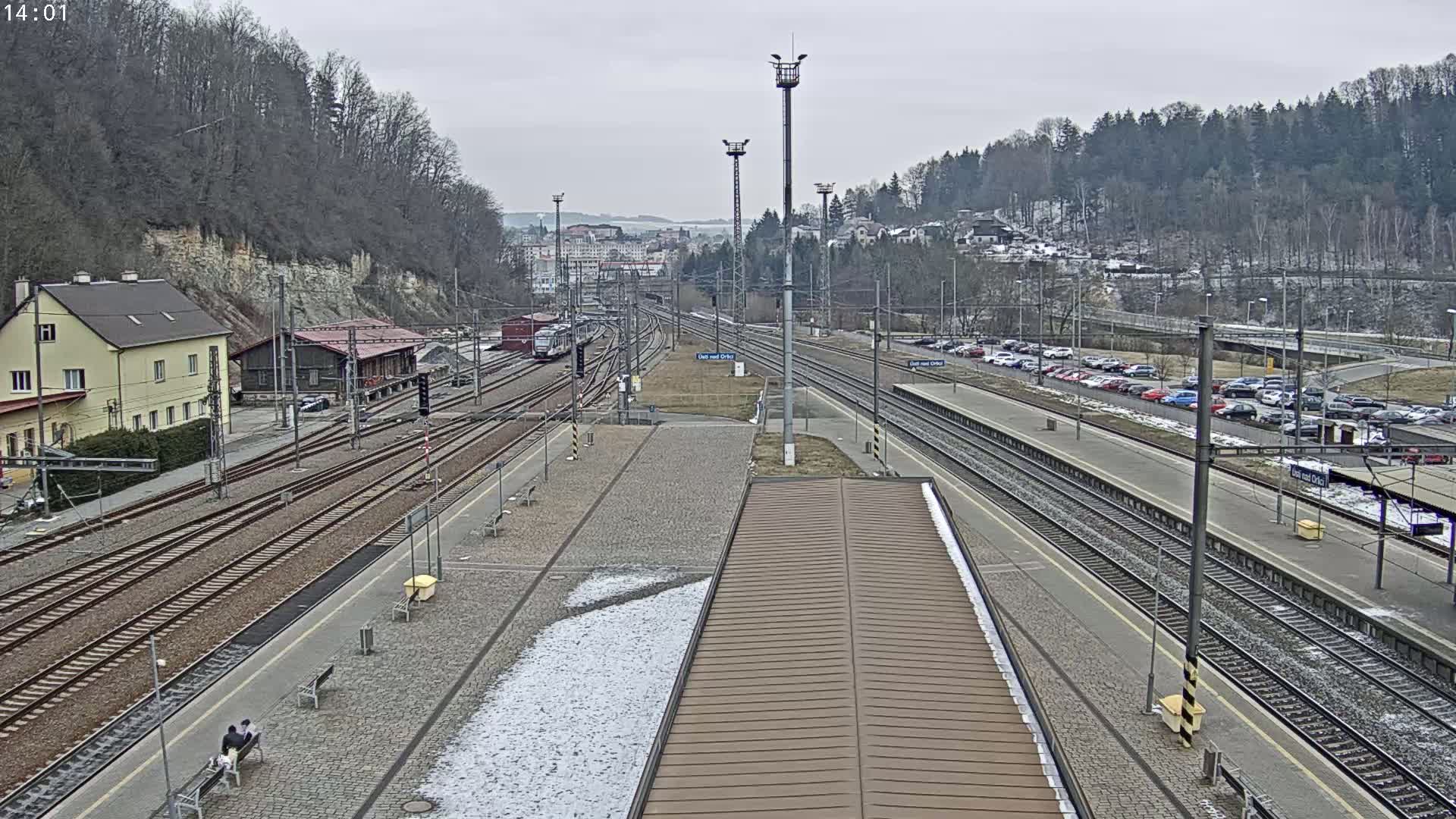 A train station with multiple tracks, a few buildings, and a parking lot is visible on an overcast day with patches of snow and ice on the ground.