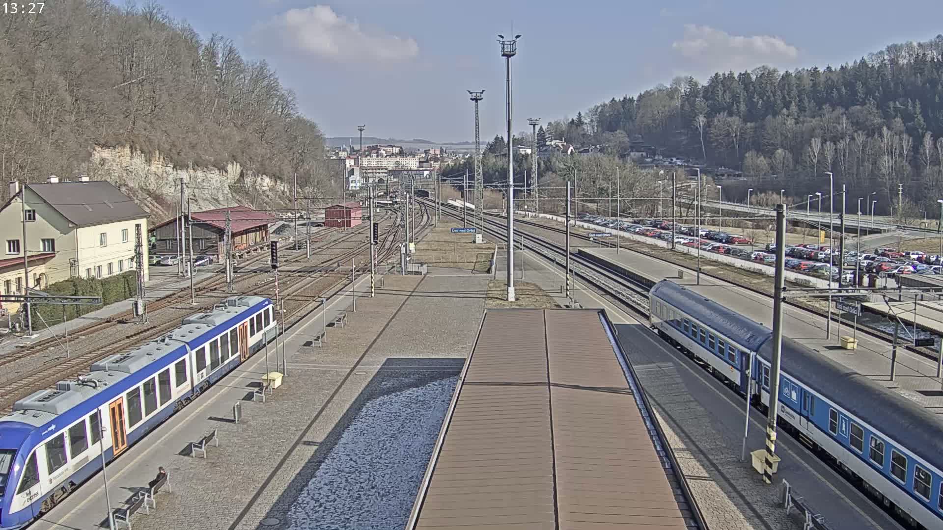 A train station on a sunny day with two trains at the platform, surrounded by buildings and trees.
