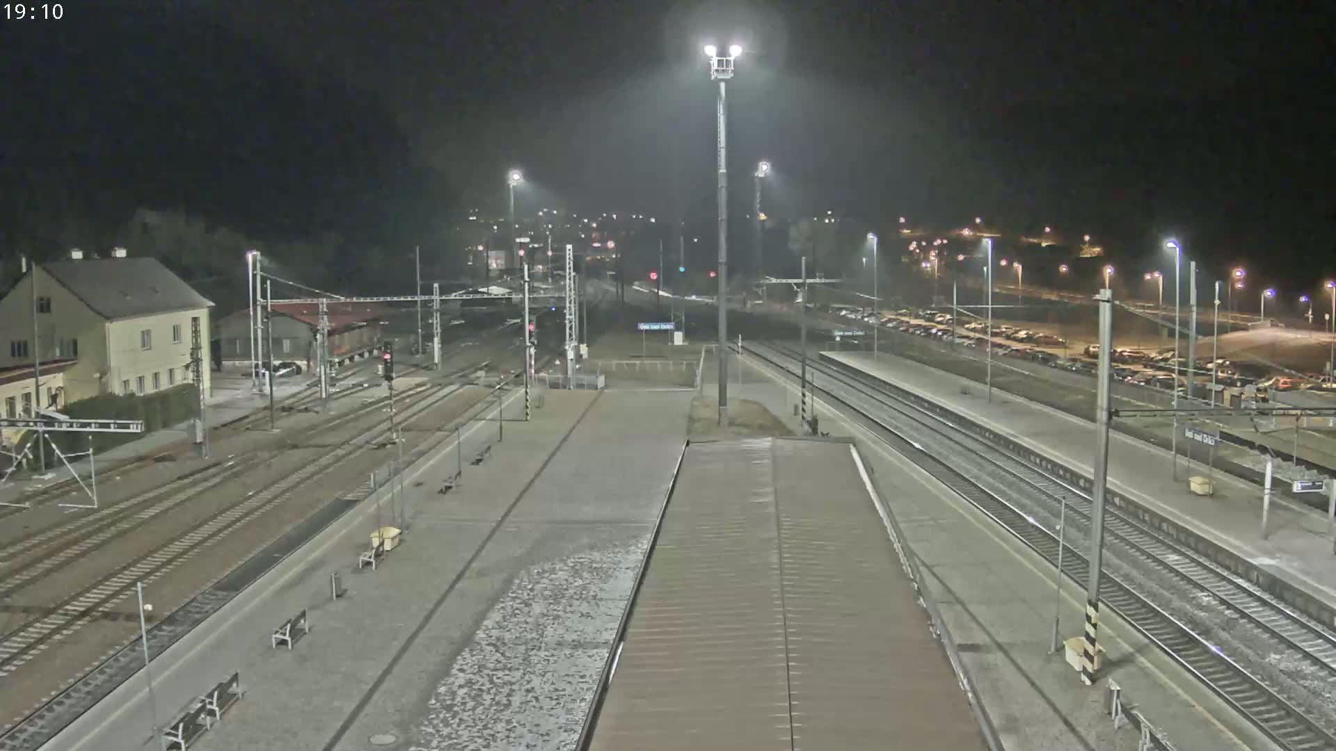 A nighttime view of a train station with multiple train tracks, platforms, and a parking lot, illuminated by bright lights.