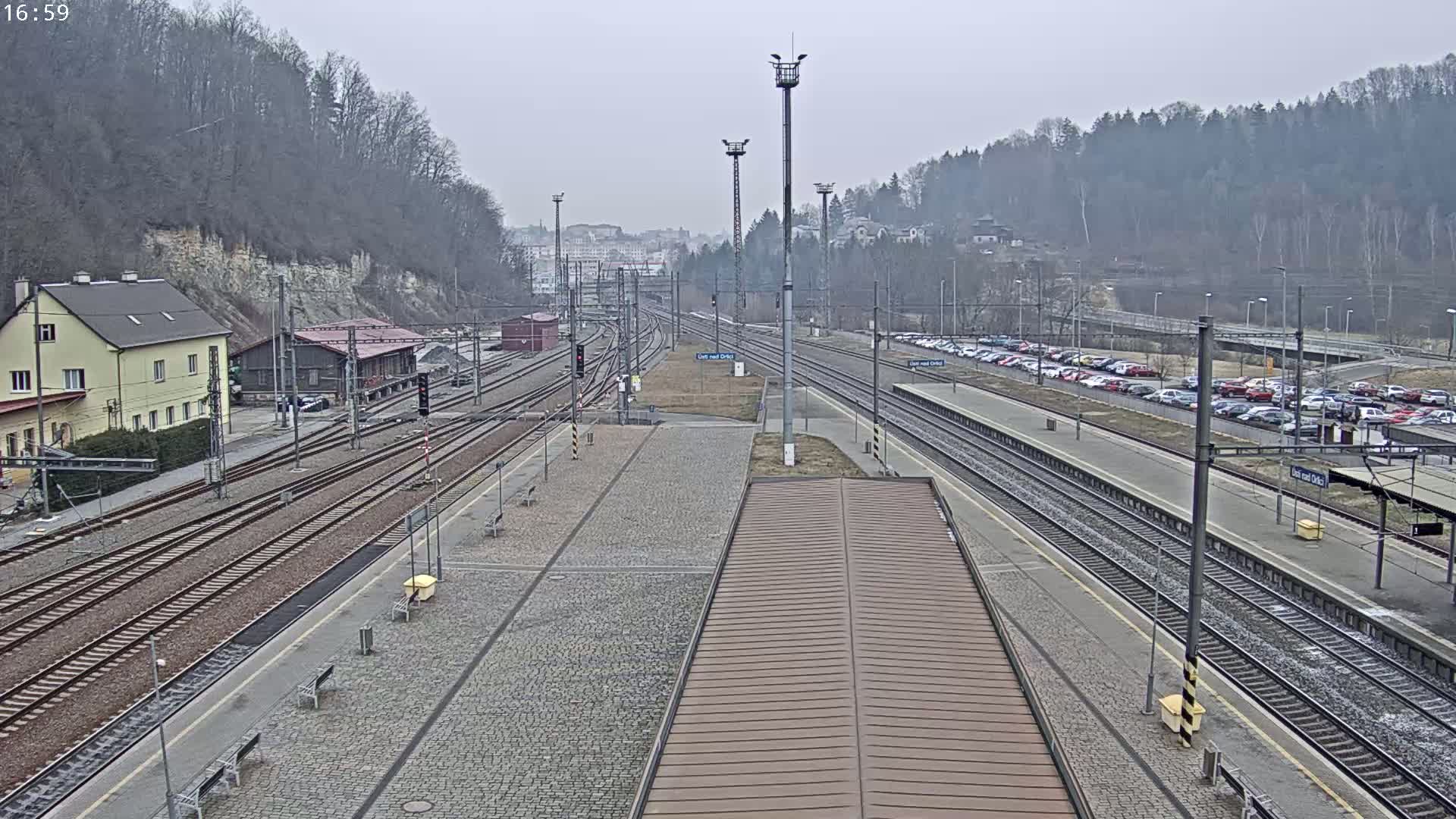 A train station with multiple tracks, a large parking lot filled with cars, and a building nestled beside a hillside under an overcast sky.