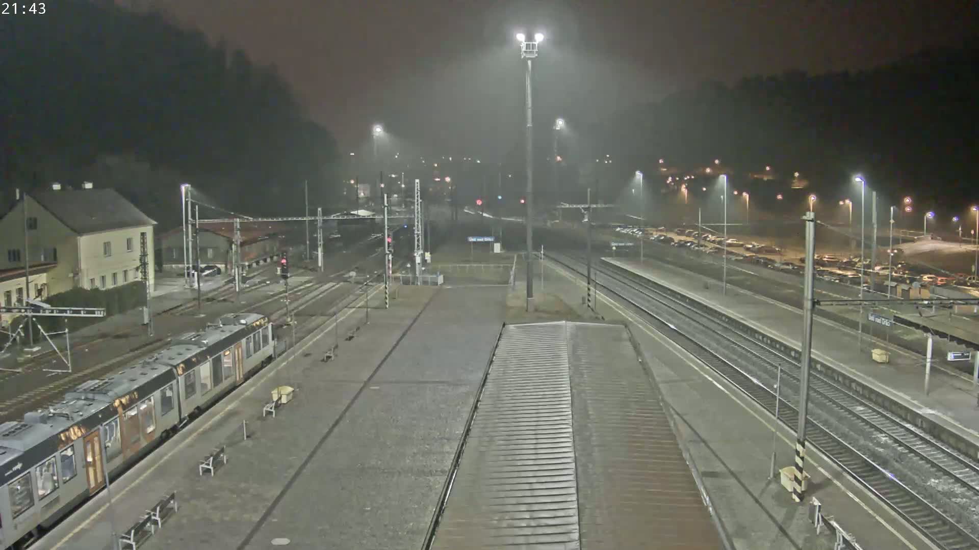 A nighttime view of a train station with a train at the platform, under misty or rainy conditions.