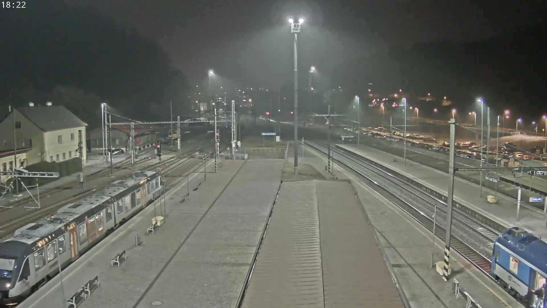 A nighttime view of a train station with two trains, one on each side of the platform, under hazy or foggy conditions.