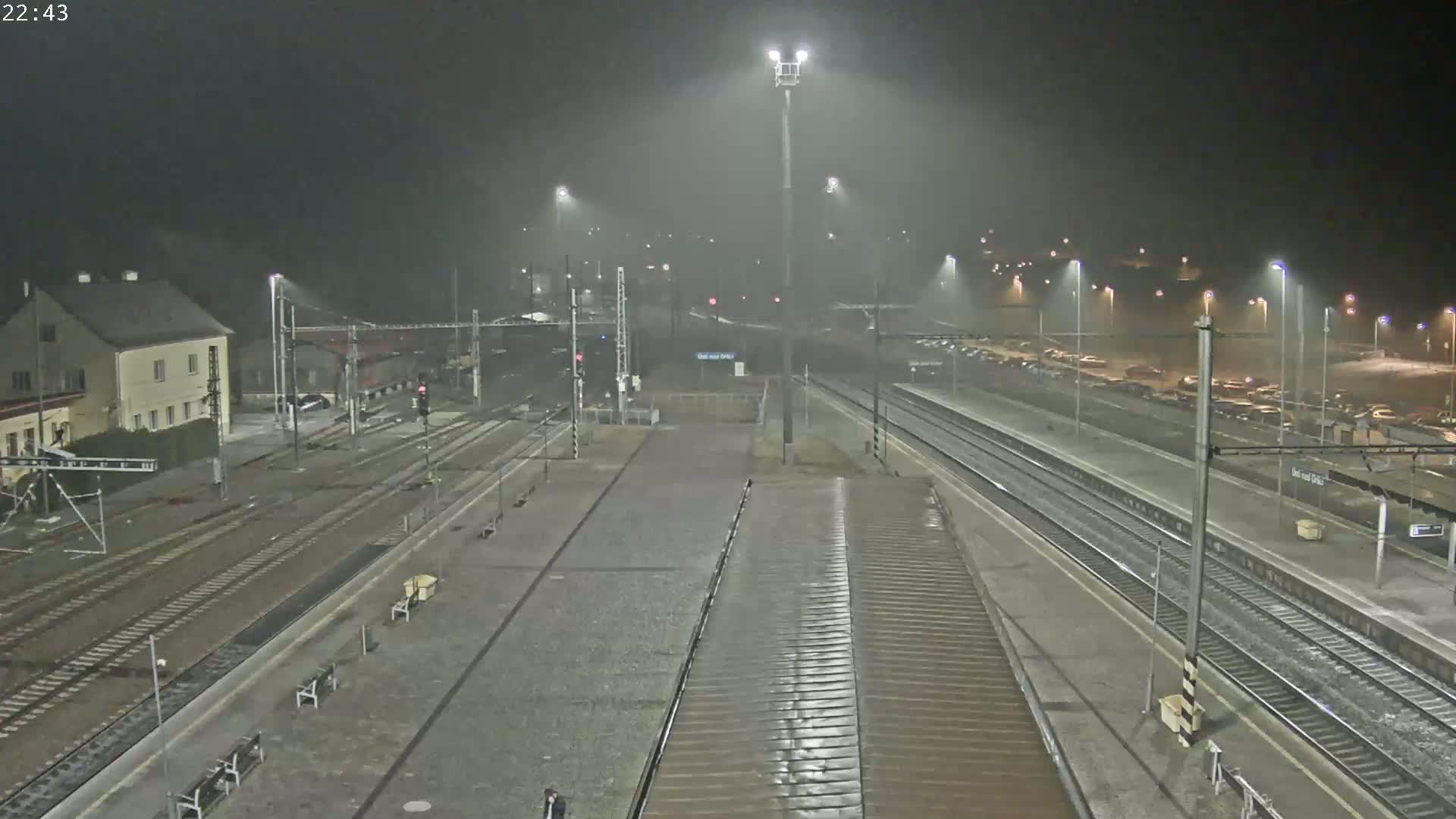 A nighttime view of a train station with multiple tracks under rainy conditions, showing a building and a parking lot in the background.