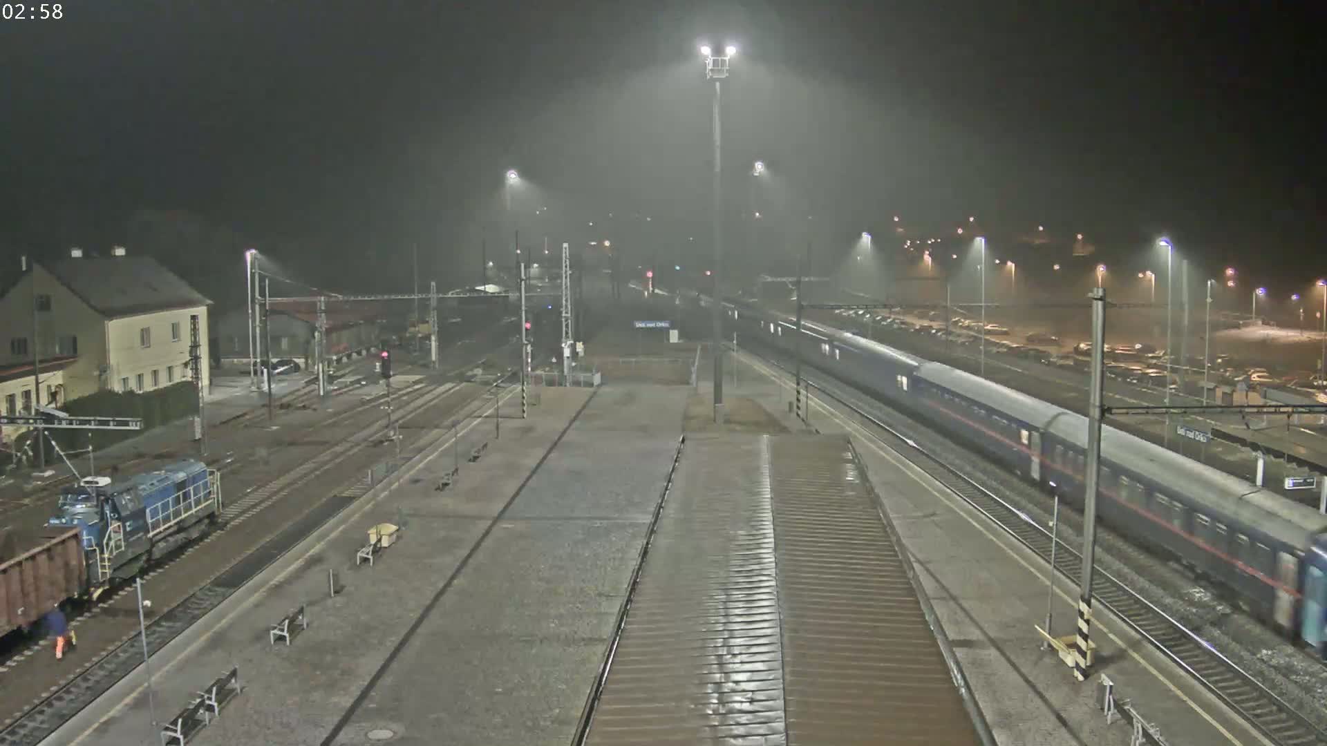 A long passenger train passes through a dimly lit train station at night in foggy or rainy conditions.