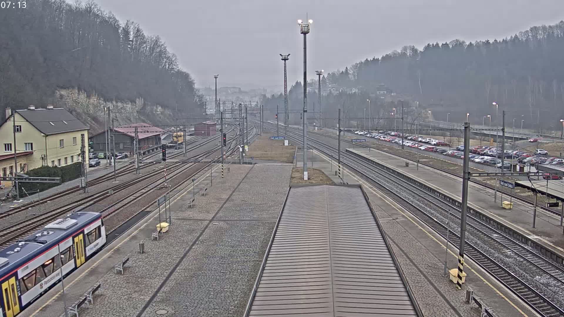 A train station on an overcast day with multiple train tracks, a train at a platform, and a parking lot filled with cars.