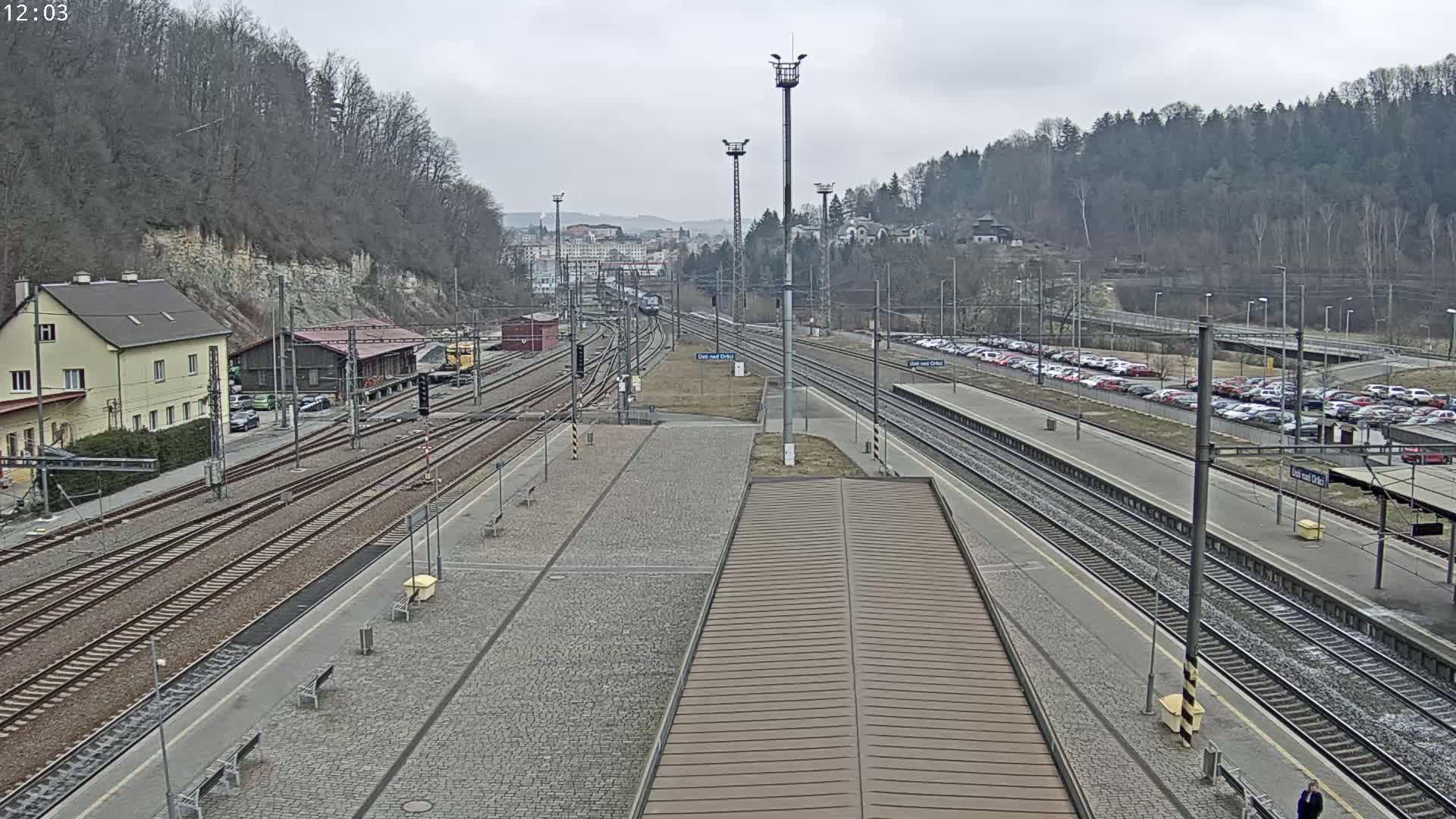 A high-angle view of a train station with multiple tracks, a large parking lot, and buildings on a cloudy day.