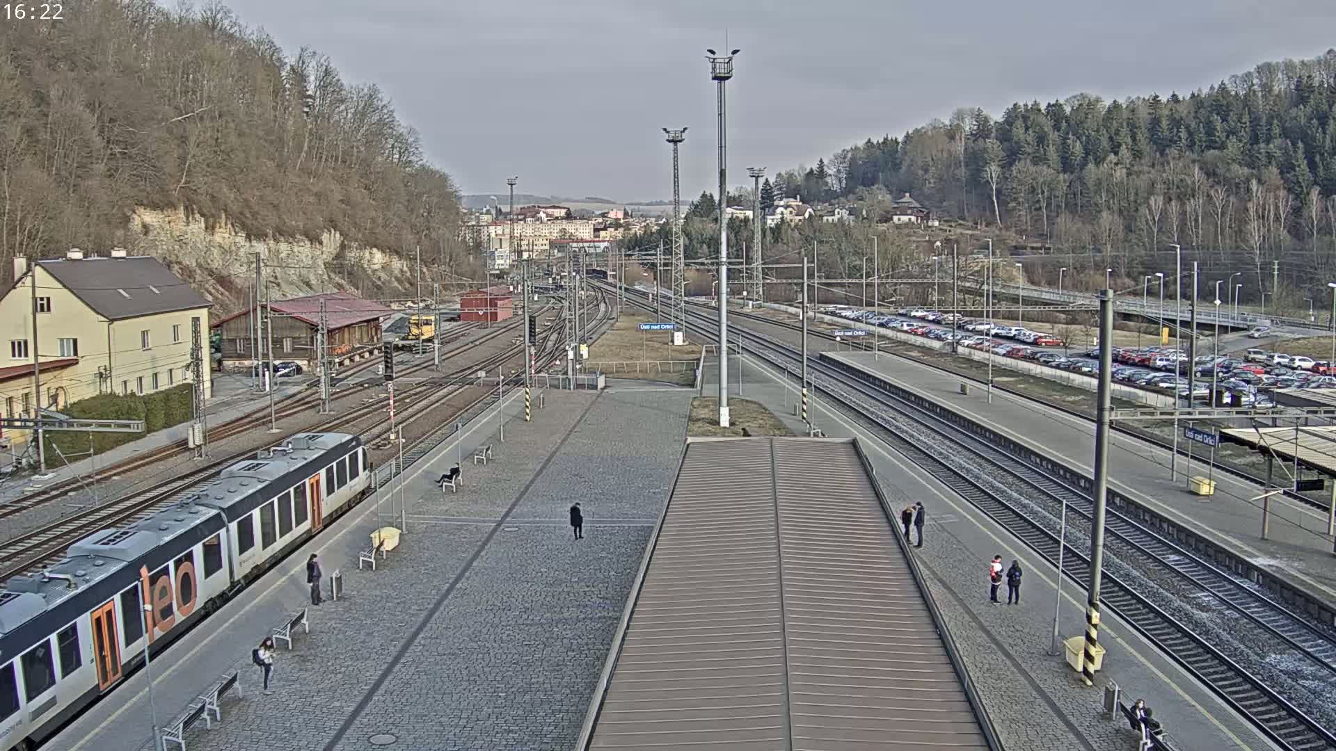 A train station with several train tracks, a passenger train, a large parking lot, and a few people standing around on a partly cloudy day.