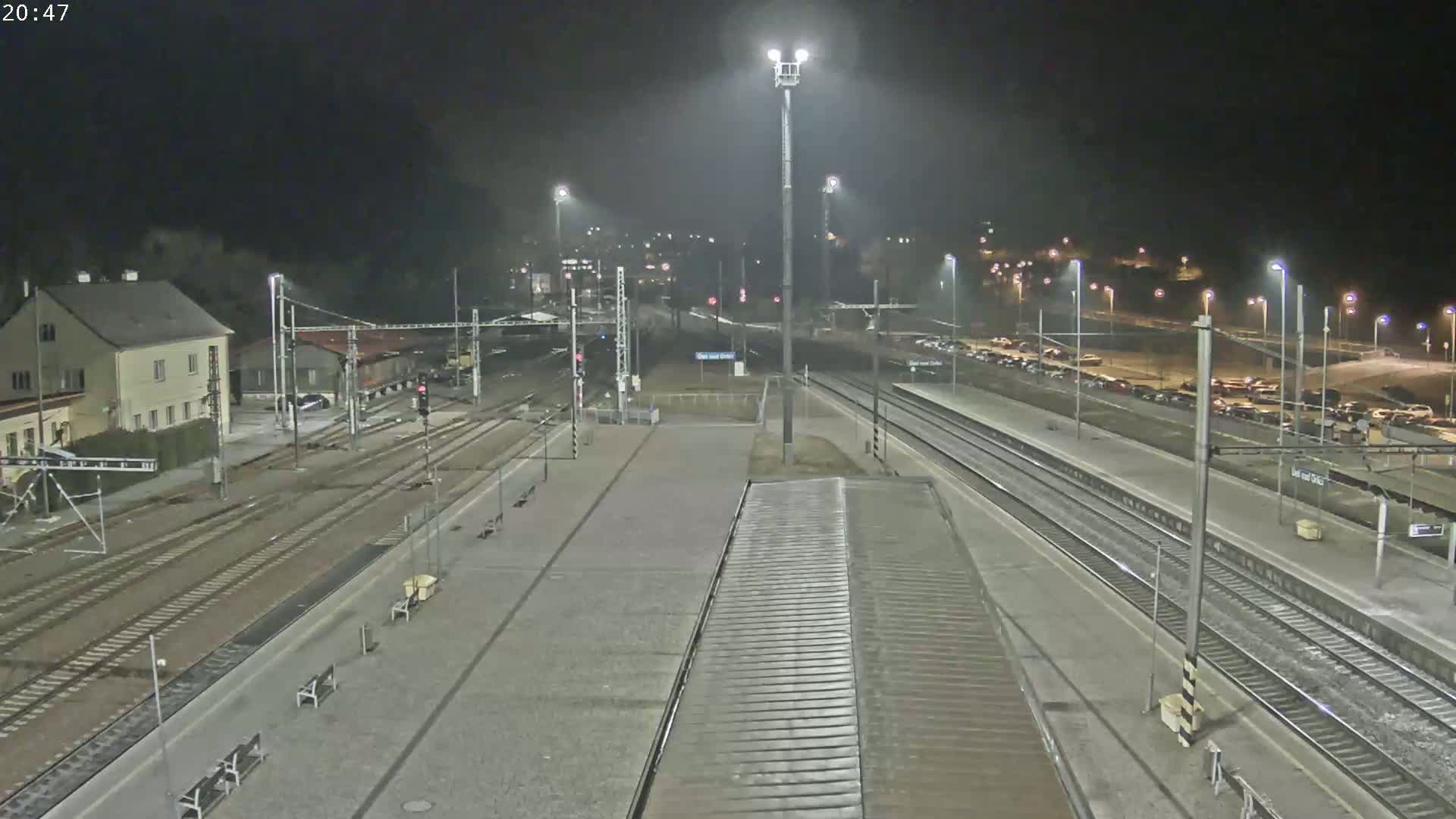 A nighttime view of a train station with multiple tracks, platforms, and a parking lot filled with cars under artificial lighting.