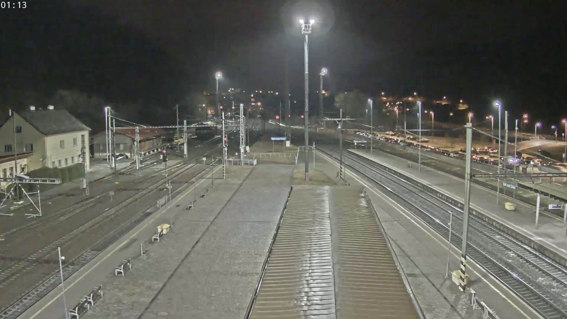 A nighttime view of a train station with multiple tracks, platforms, and a parking lot filled with cars, under dark and clear skies.
