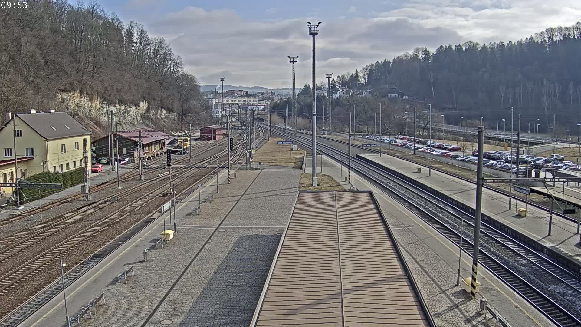 A train station with multiple tracks, a parking lot, and a building situated on a hillside under a partly sunny sky.