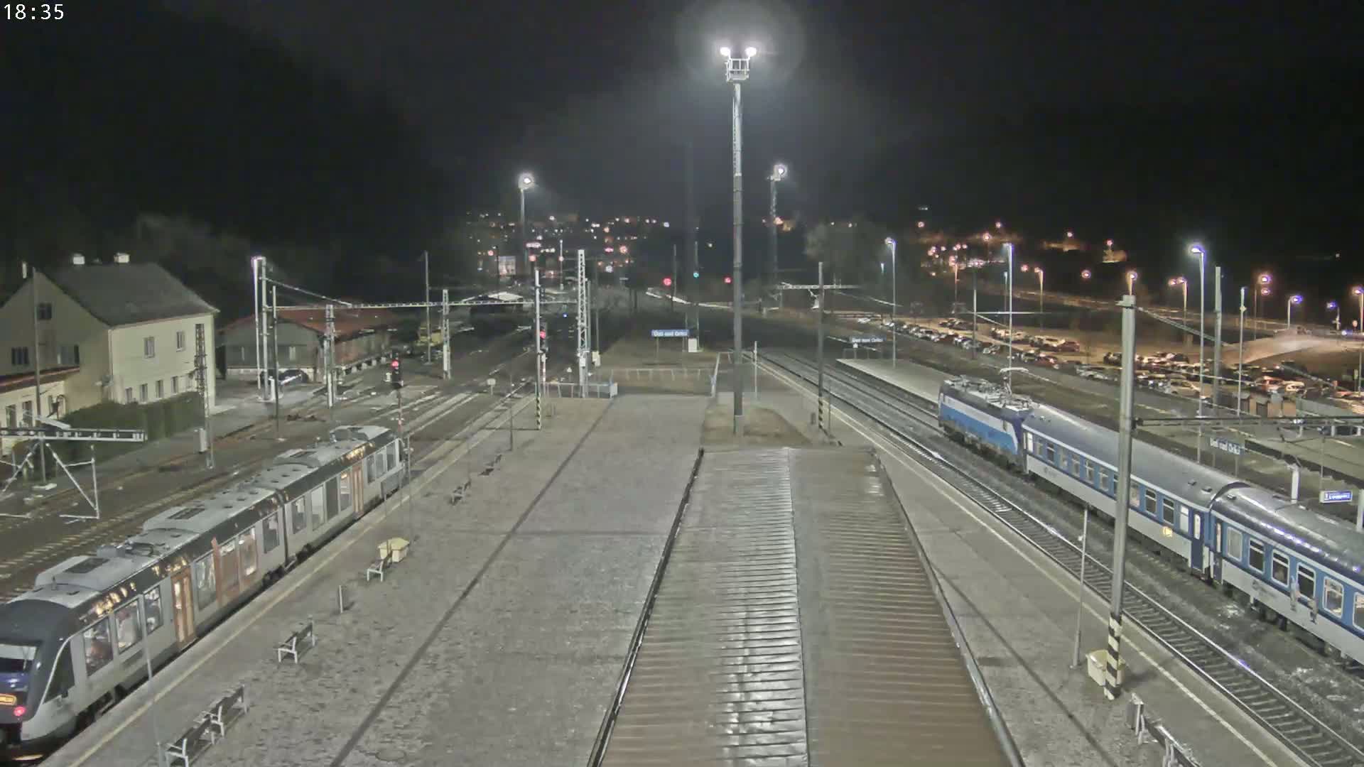 At night, two trains are at a station platform, with a large, empty parking lot visible in the background.