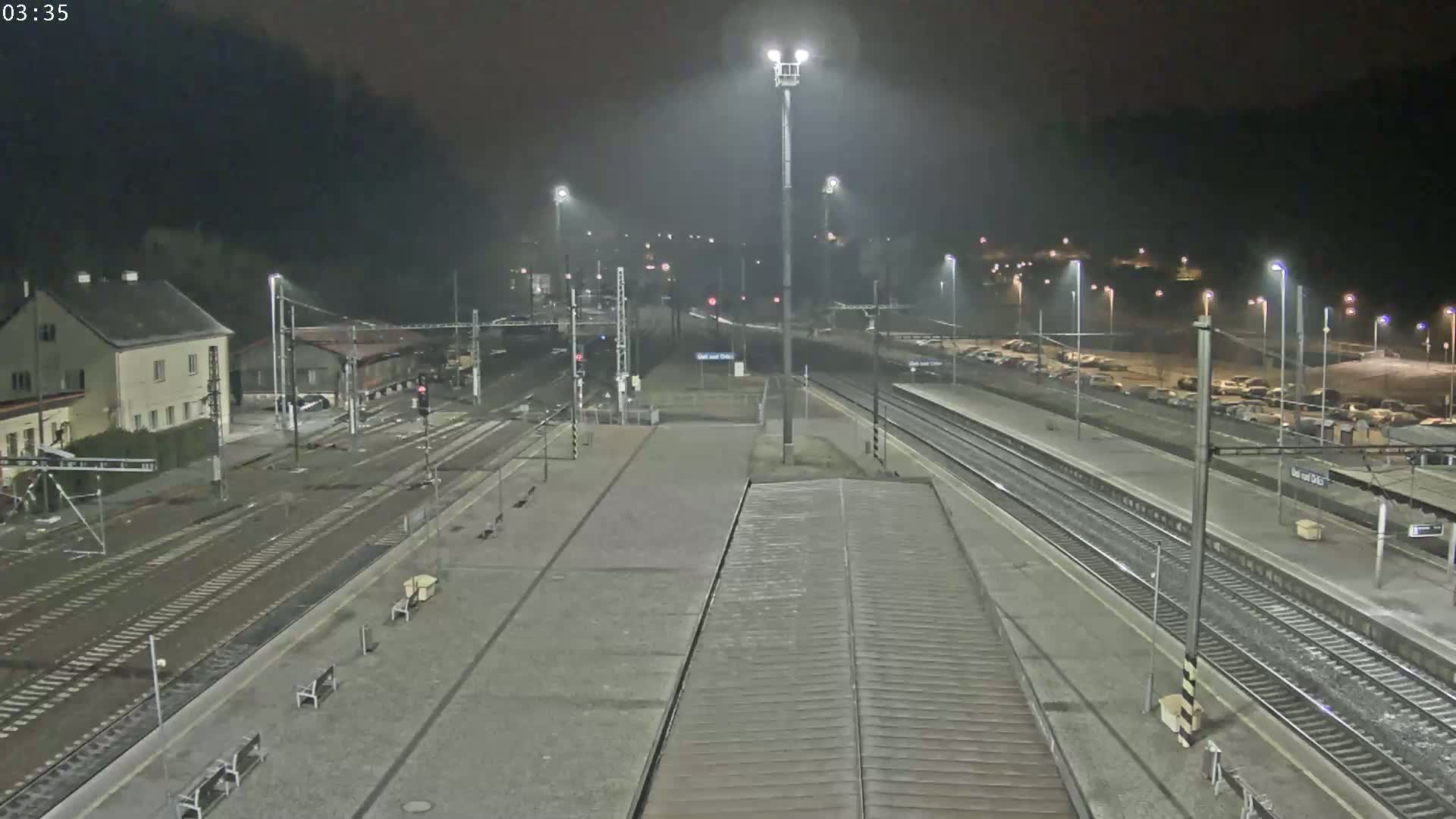 A nighttime view of a train station with multiple train tracks, a building, and a parking lot, under hazy or foggy conditions.