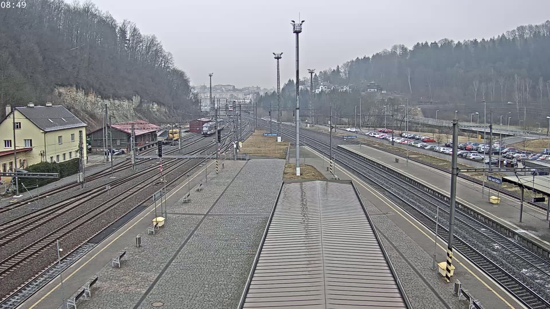 A train station with multiple tracks, a few trains, and a parking lot is seen on an overcast day.