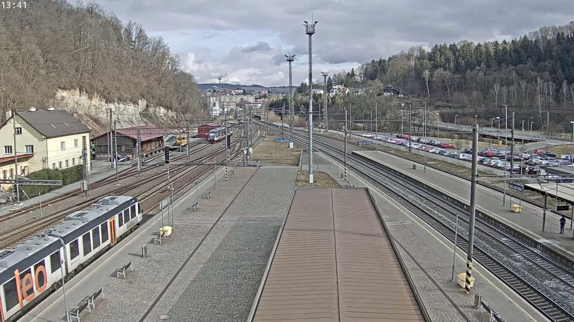 A train station with multiple train tracks, a passenger train, several buildings, and a large parking lot, all under a cloudy sky.
