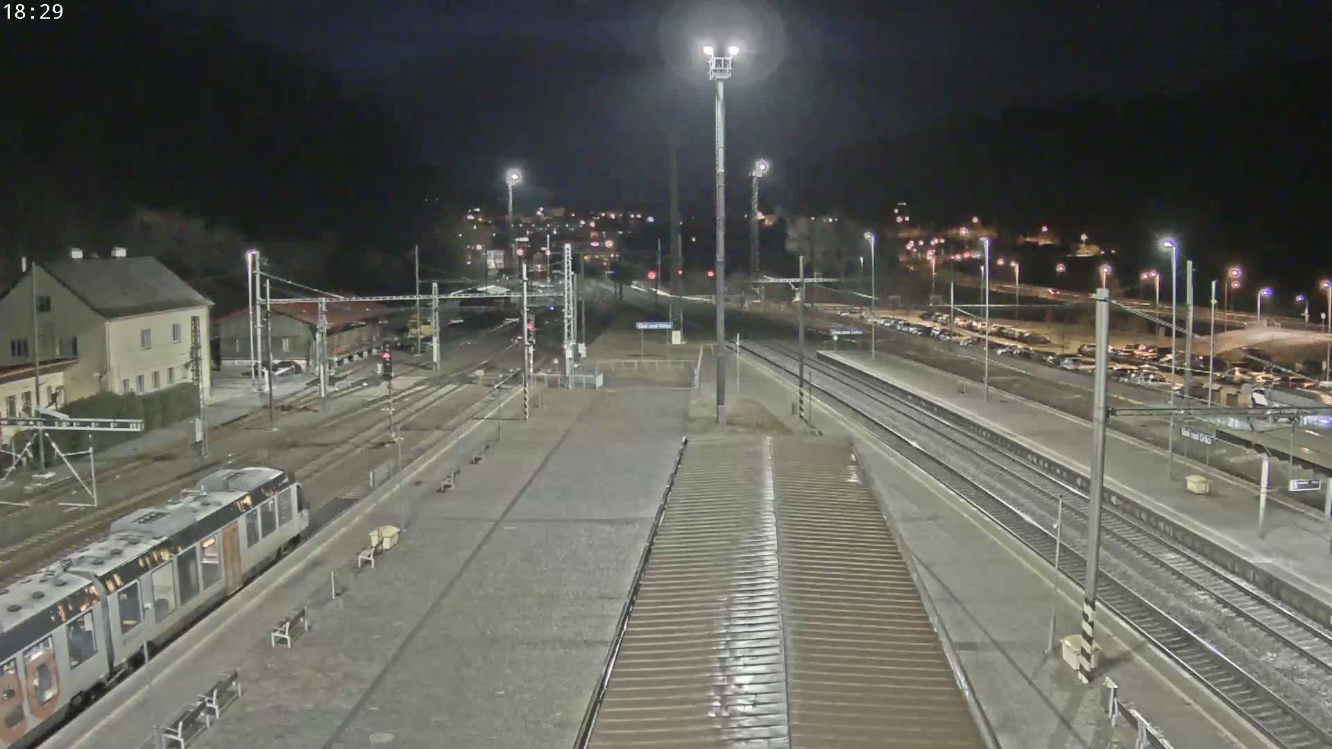 A nighttime view of a train station with a train at the platform, multiple tracks, and a parking lot visible in the background under clear skies.