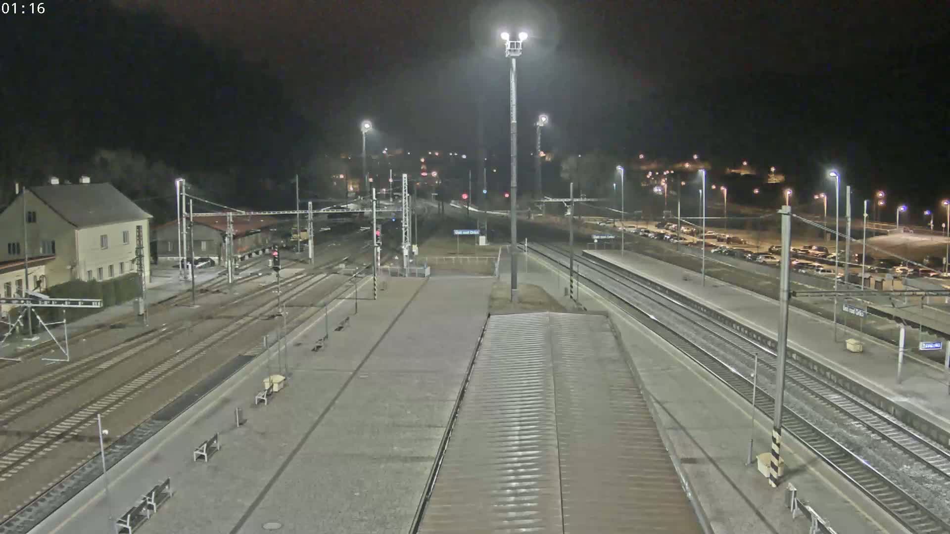 A nighttime view of a train station with multiple tracks, platforms, and a parking lot, illuminated by artificial lights.