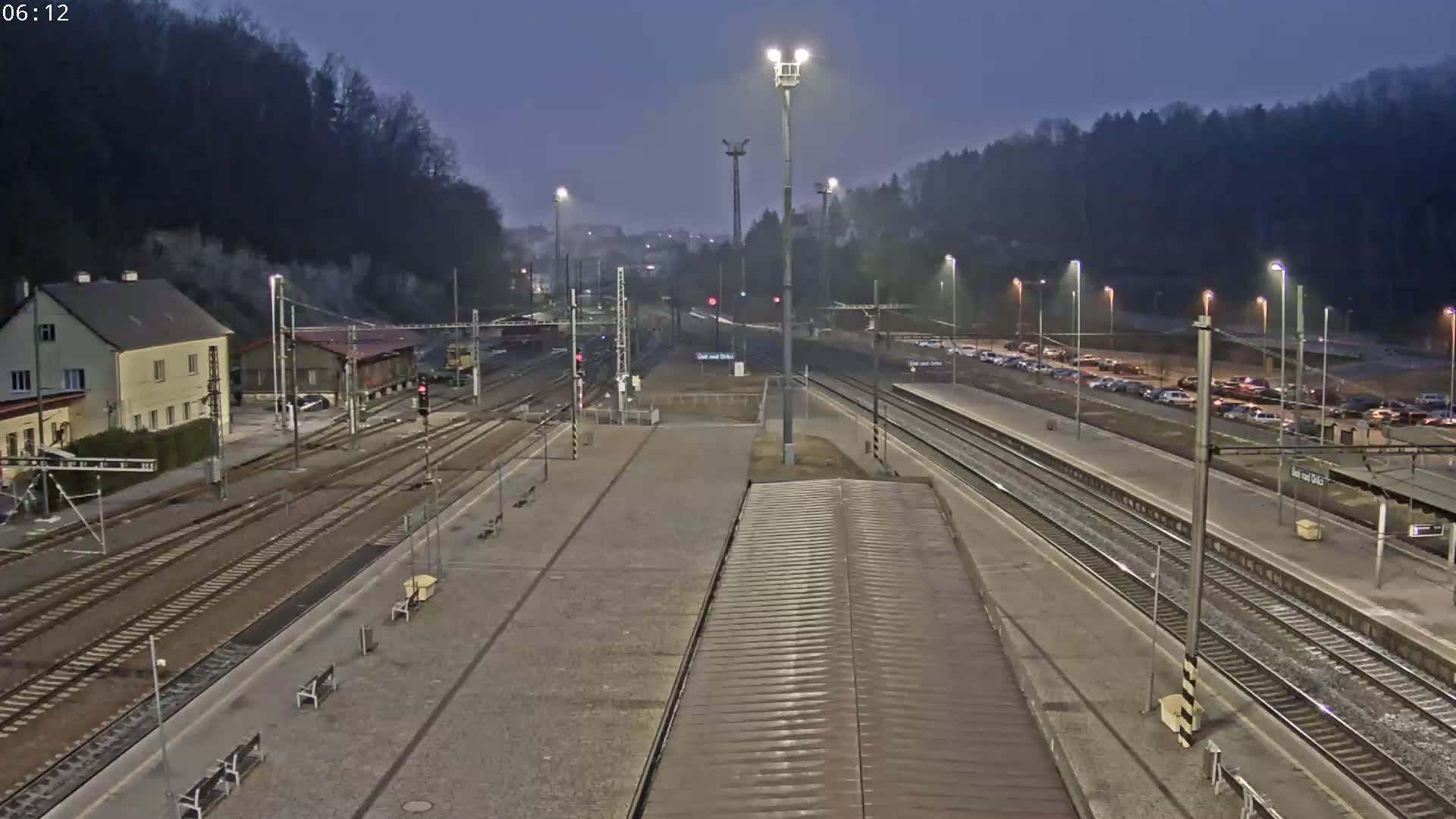 A nighttime view of a train station with multiple train tracks, a building, and several cars parked nearby under a twilight sky.