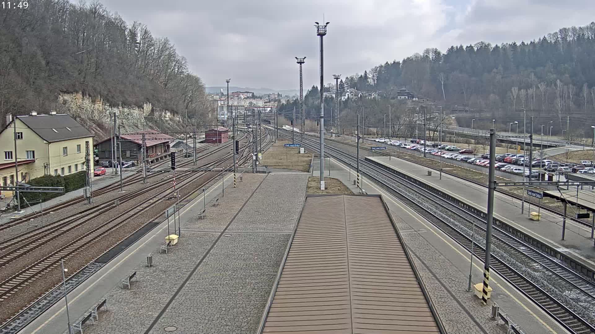 A train station with multiple tracks and a parking lot is visible under an overcast sky.