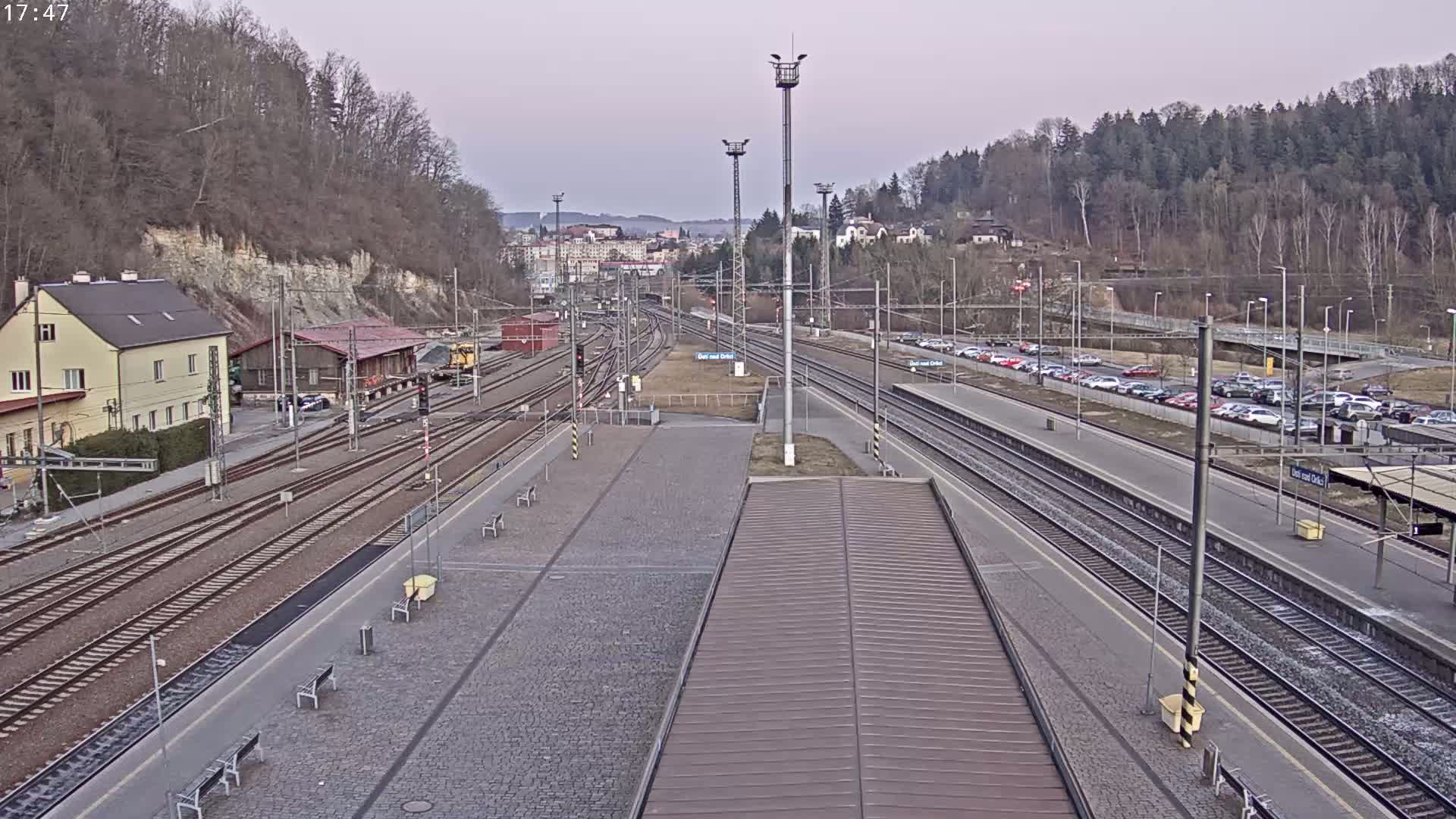 A train station with multiple tracks, a building, and a parking lot is seen on an overcast day.