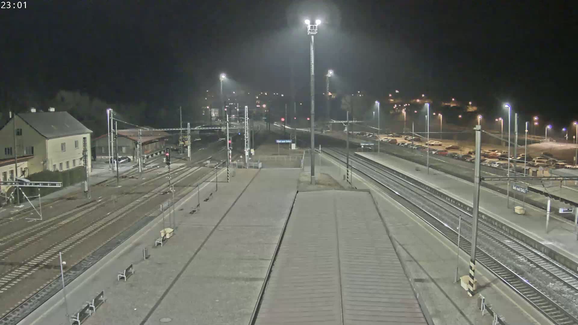 A nighttime view of a train station with multiple train tracks, a building, and a parking lot filled with cars under bright artificial lights.