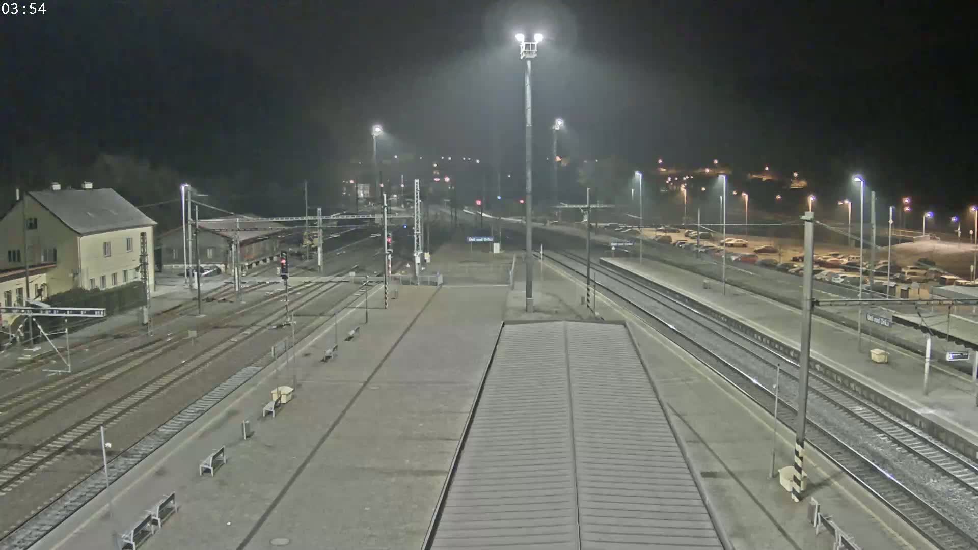A nighttime view of a train station with multiple tracks, a parking lot filled with cars, and a building in the background under a foggy sky.