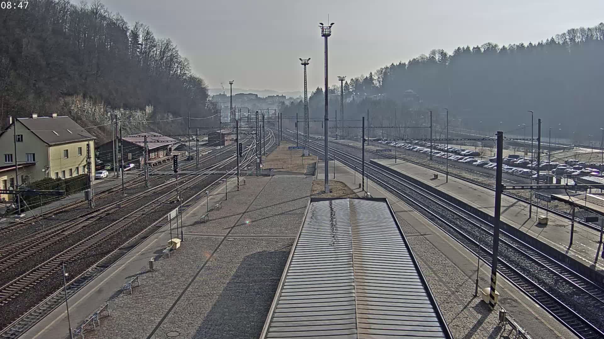 A high-angle view of a train station on a sunny day, showing multiple train tracks, a parking lot filled with cars, and buildings near a wooded hillside.