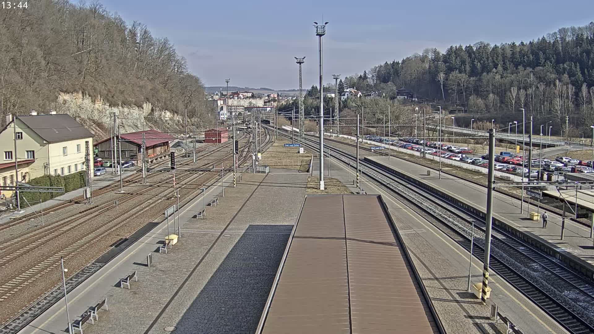 A train station with multiple tracks and a large parking lot is visible on a sunny day, surrounded by trees and hills.