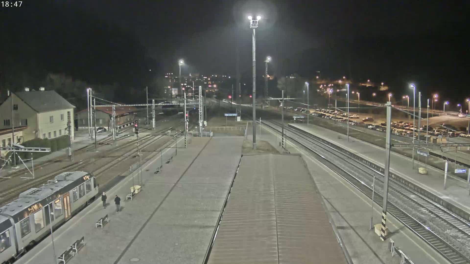 A nighttime view of a train station with a train on the platform, several parked cars in a lot, and a building in the background under clear skies.
