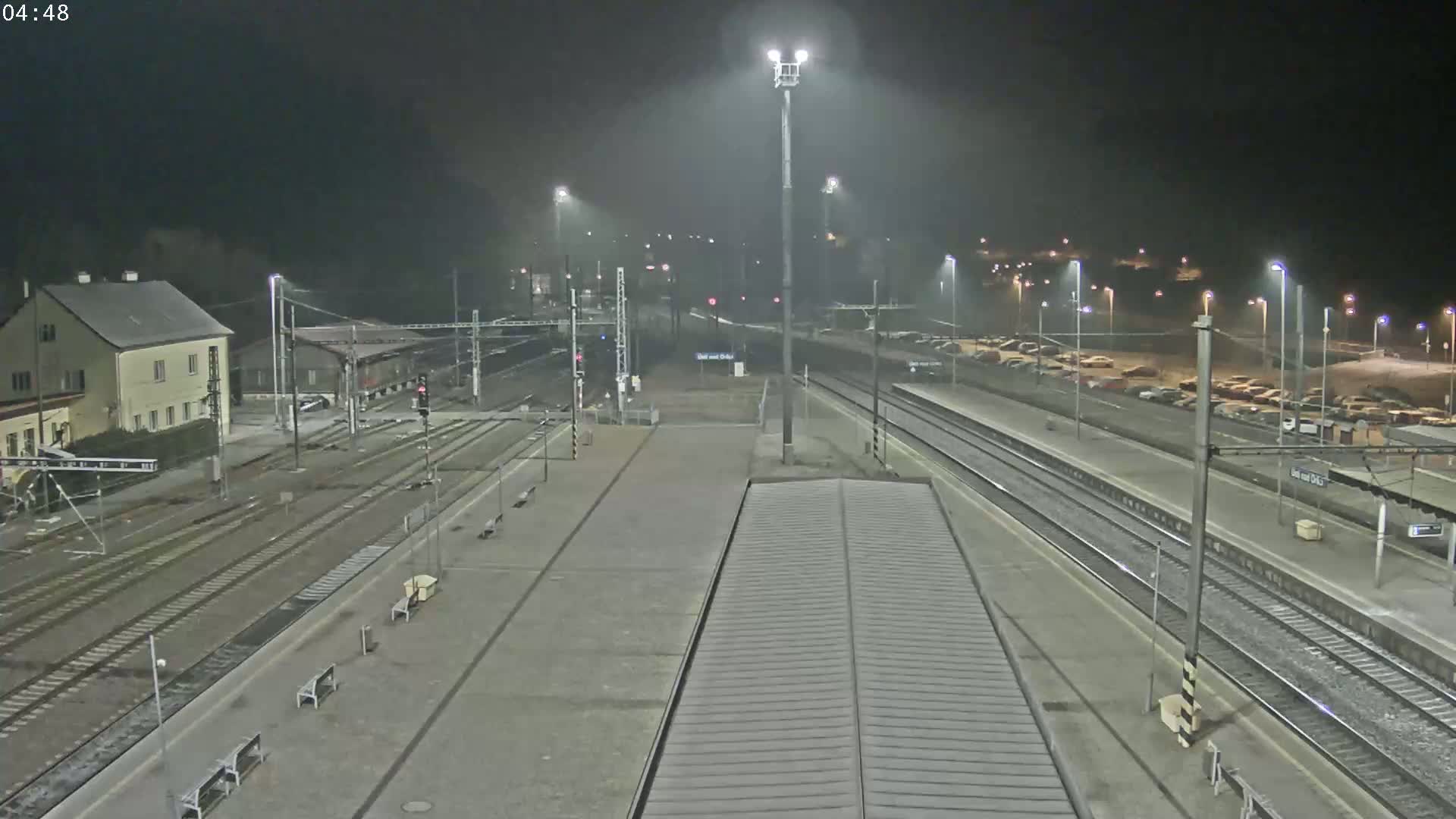 A nighttime view of a train station with multiple tracks, a parking lot, and a building under hazy or foggy conditions.