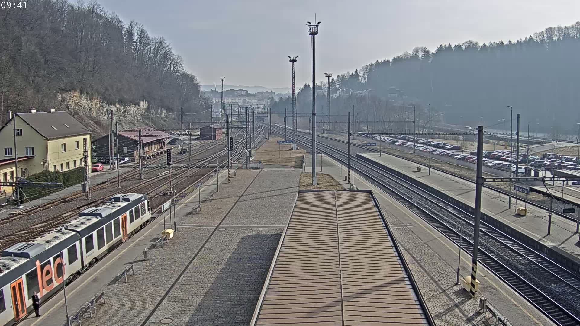 A train station with multiple train tracks, a passenger train, several buildings, and a large parking lot is visible on a sunny, clear day.