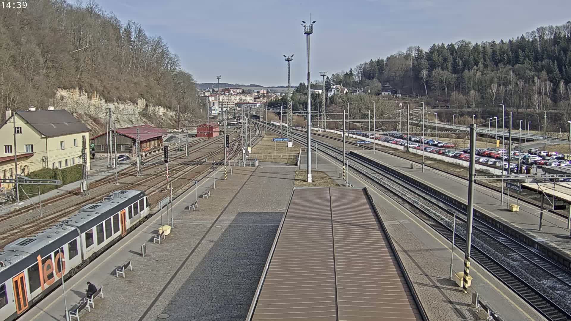 A train sits at a station platform on a sunny day, with multiple train tracks extending into the distance, alongside a parking lot filled with cars, and a hillside covered in leafless trees in the background.