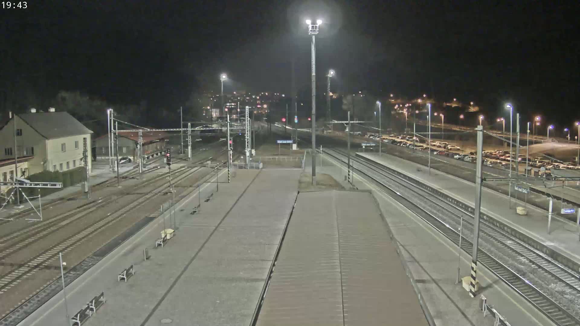 An elevated nighttime view of a train station platform with multiple train tracks, a parking lot filled with cars, and a building in the background under clear skies.