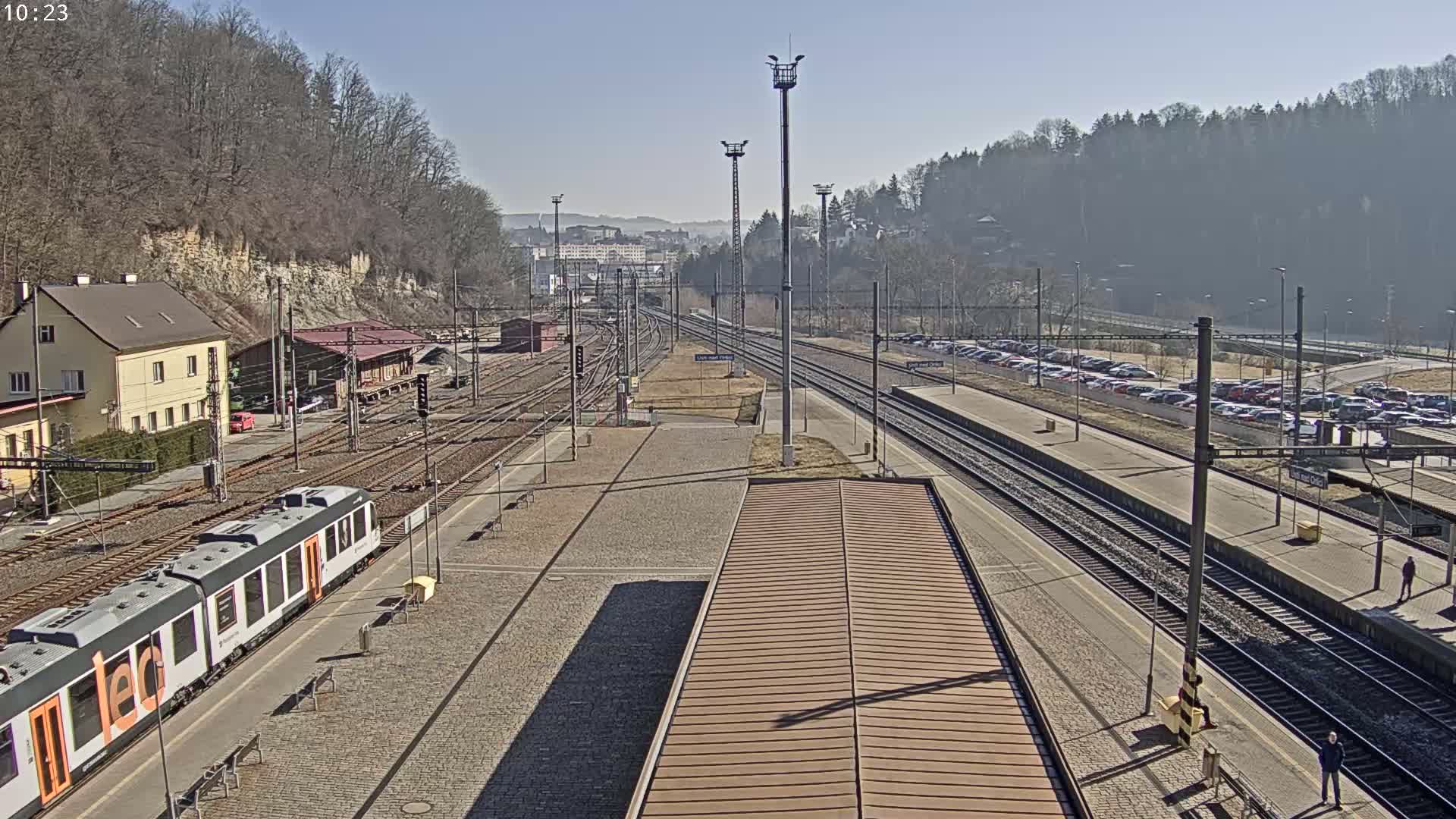 A train station on a sunny day with several train tracks, a train at the platform, and a large parking lot filled with cars.