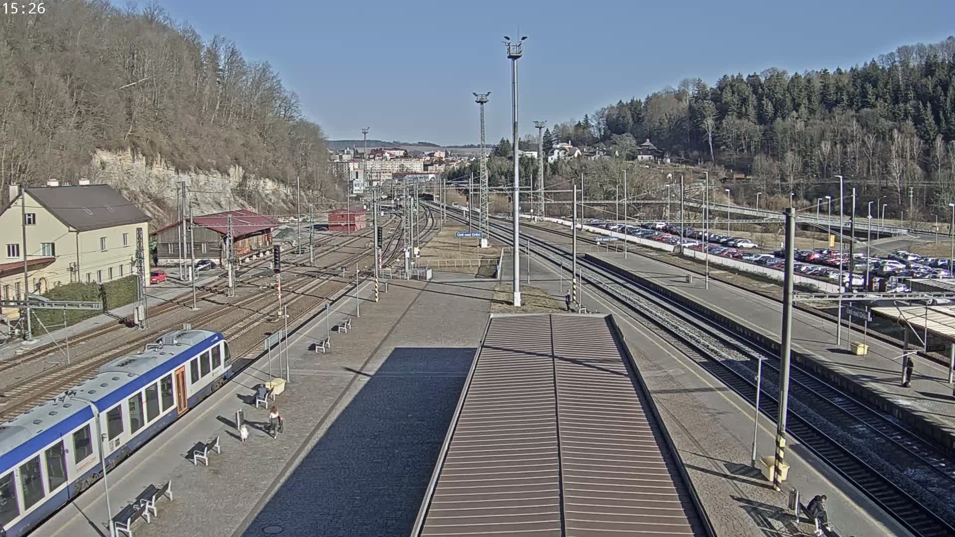 A train station on a sunny day with a train at the platform, multiple train tracks, and a large parking lot filled with cars.