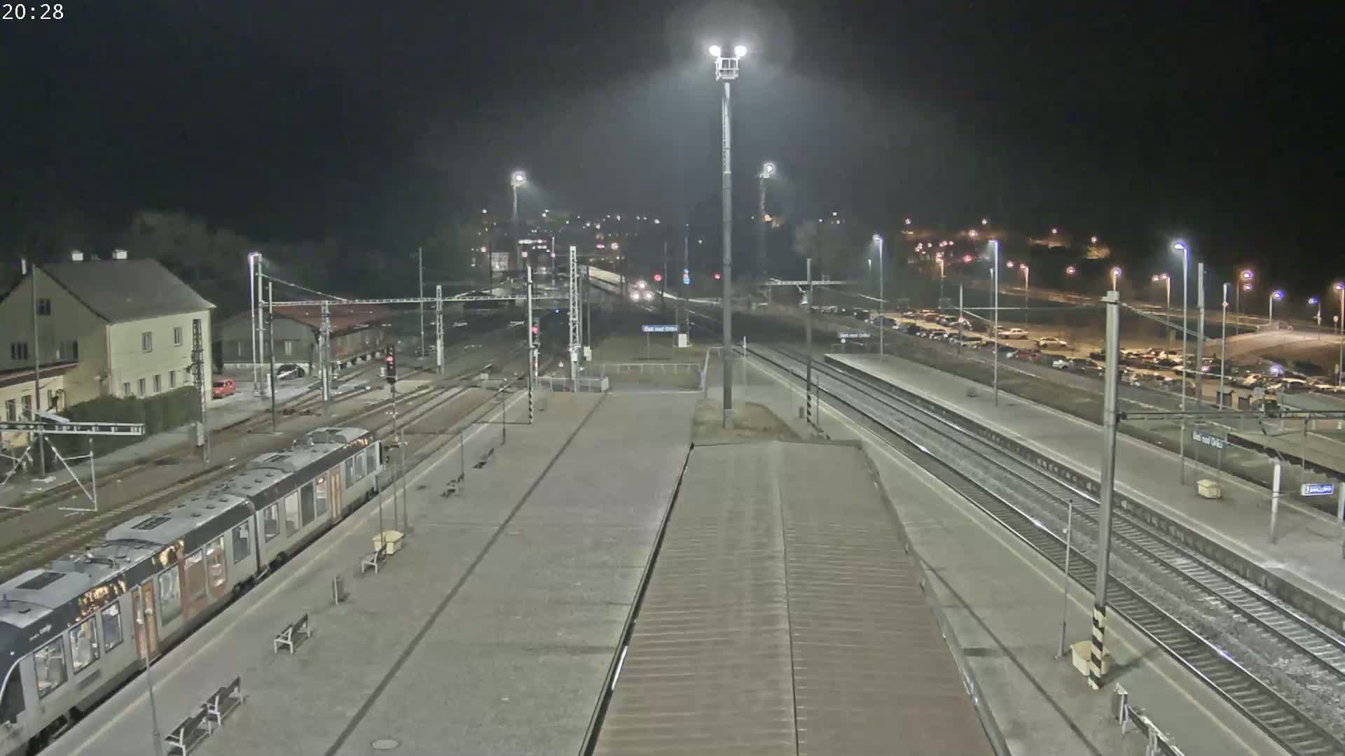 A nighttime view of a train station with a passenger train at the platform, multiple train tracks, and a parking lot with cars visible in the distance under partly cloudy skies.