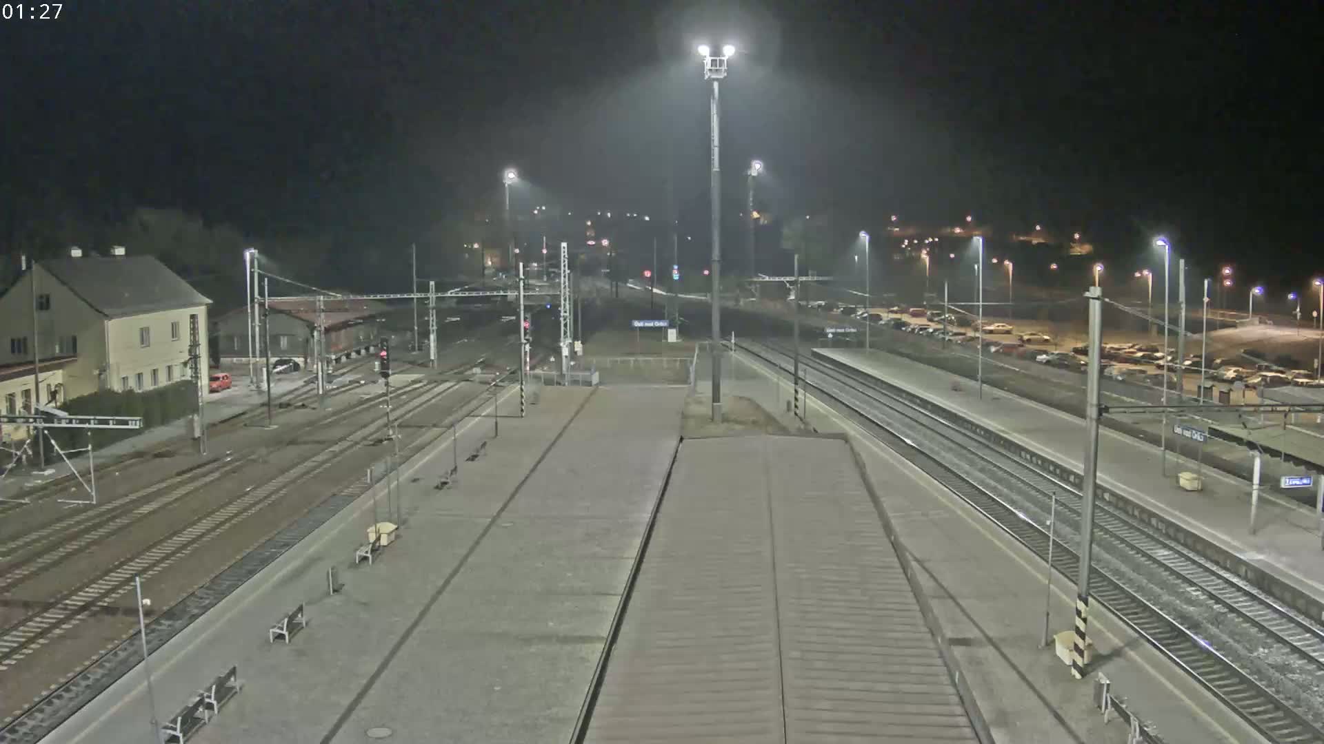 A nighttime view of a train station with multiple tracks, platforms, a parking lot, and a building, under slightly hazy conditions.