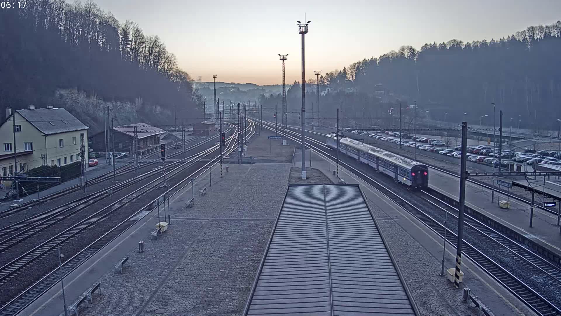 A passenger train is departing a railway station at dawn, under a hazy sky, with a large parking lot visible nearby.