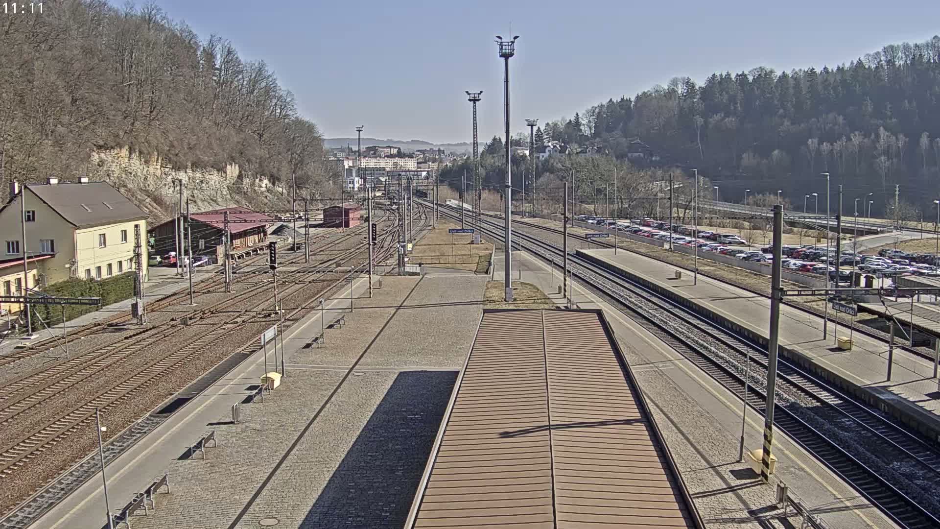 A sunny view of a train station with multiple tracks, a building, and a large parking lot filled with cars.