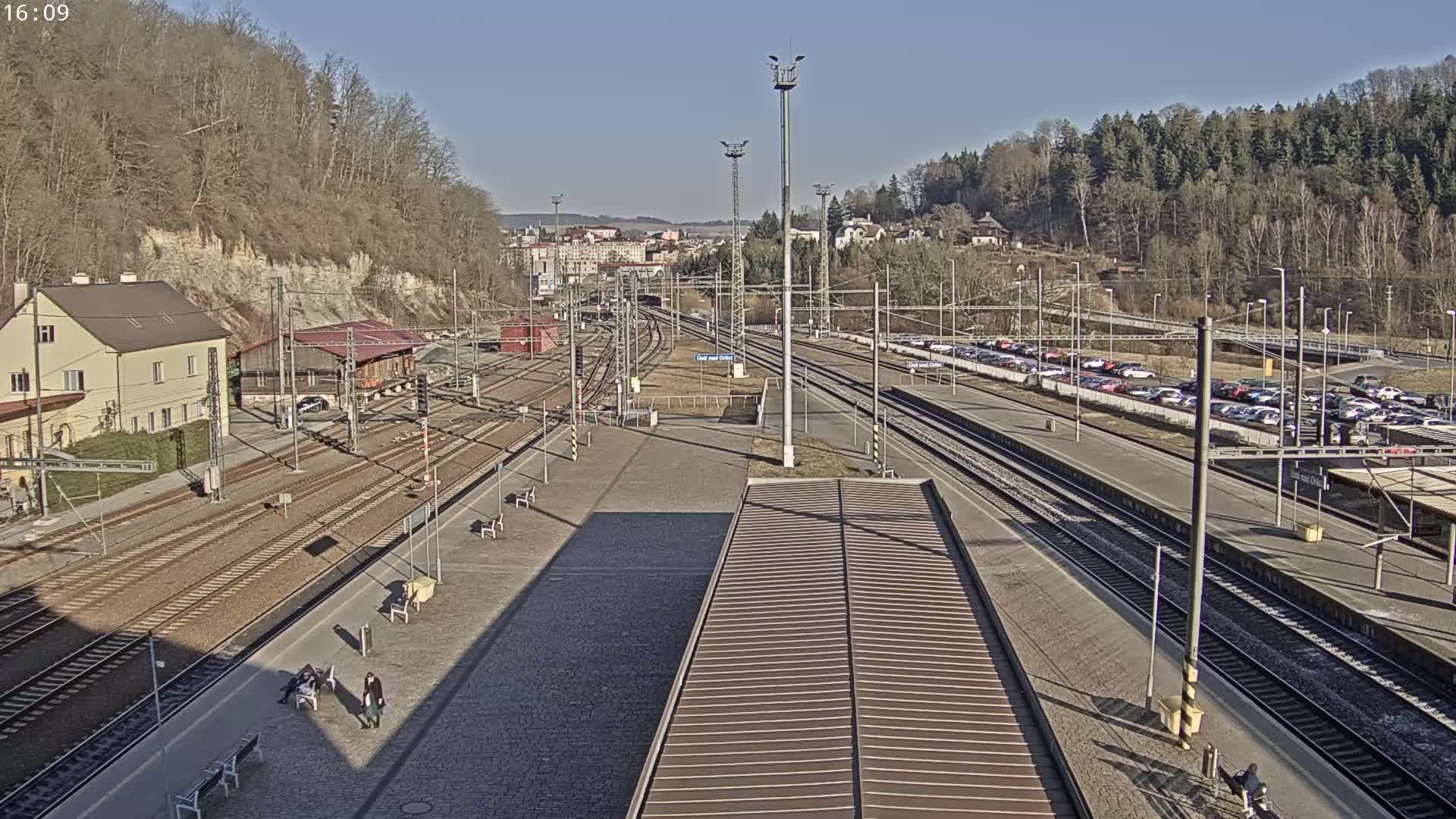 A high-angle view of a train station on a sunny day, showing multiple train tracks, a parking lot filled with cars, and a few people sitting on benches.